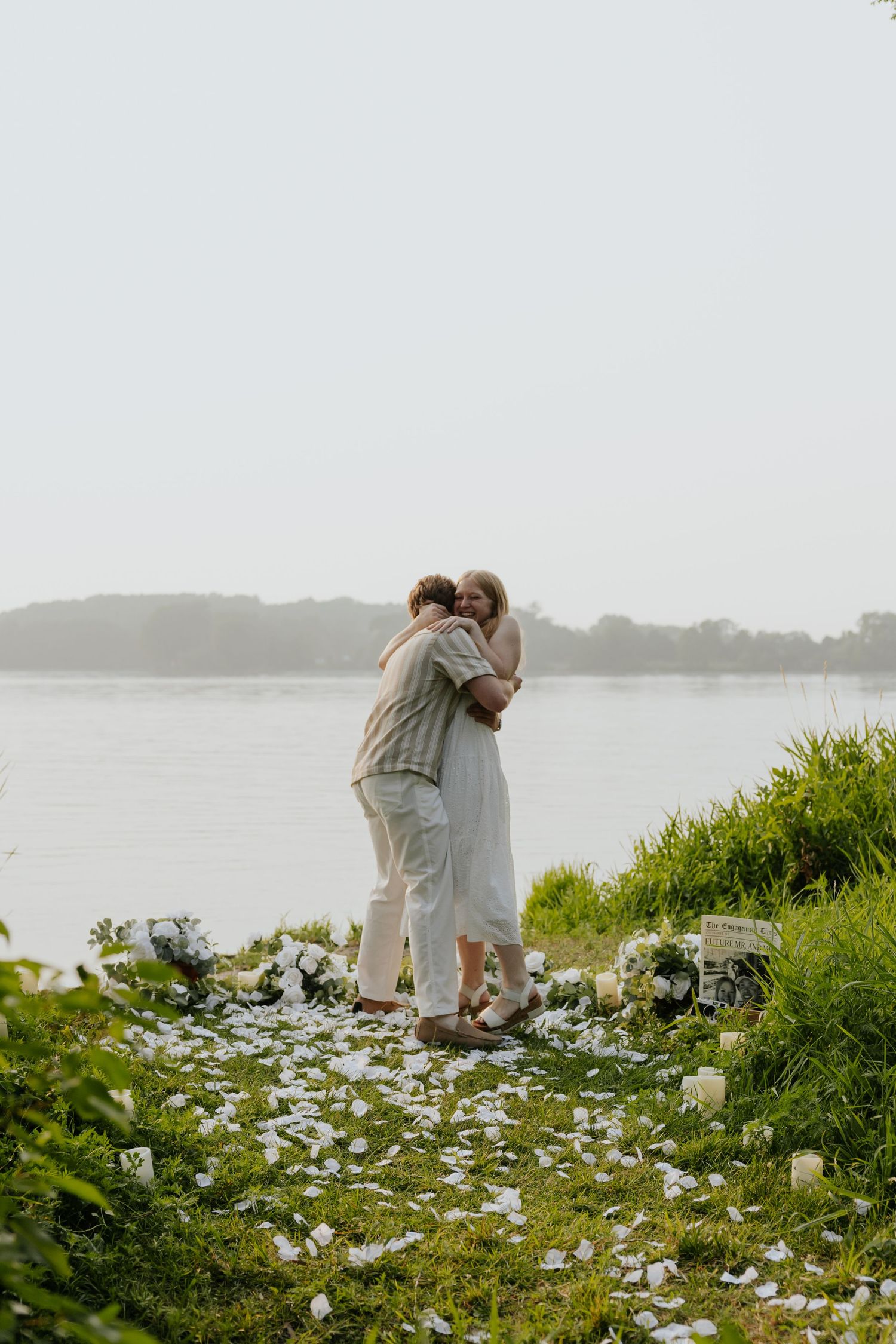 Couple embracing during a romantic lakeside proposal with white flower petals scattered on grass.