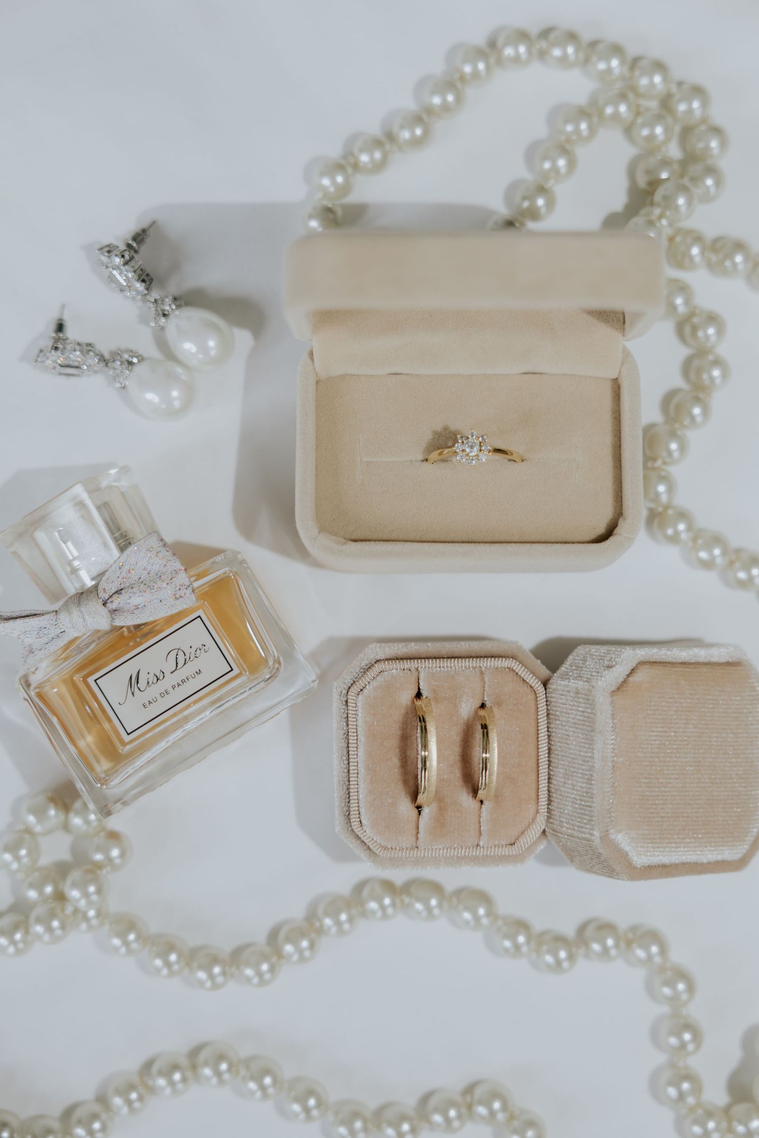 Flat lay of wedding rings in velvet boxes with Miss Dior perfume, pearl necklace, and drop earrings.