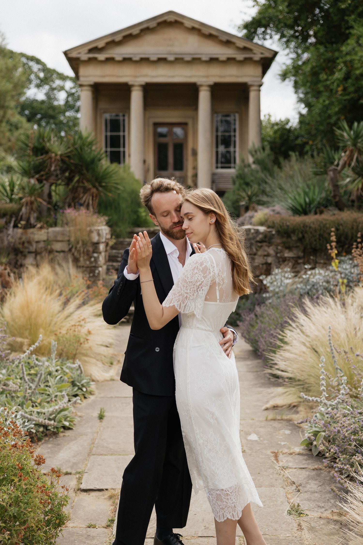 Bride and groom dancing near a neoclassical stone temple in a lush botanical garden.