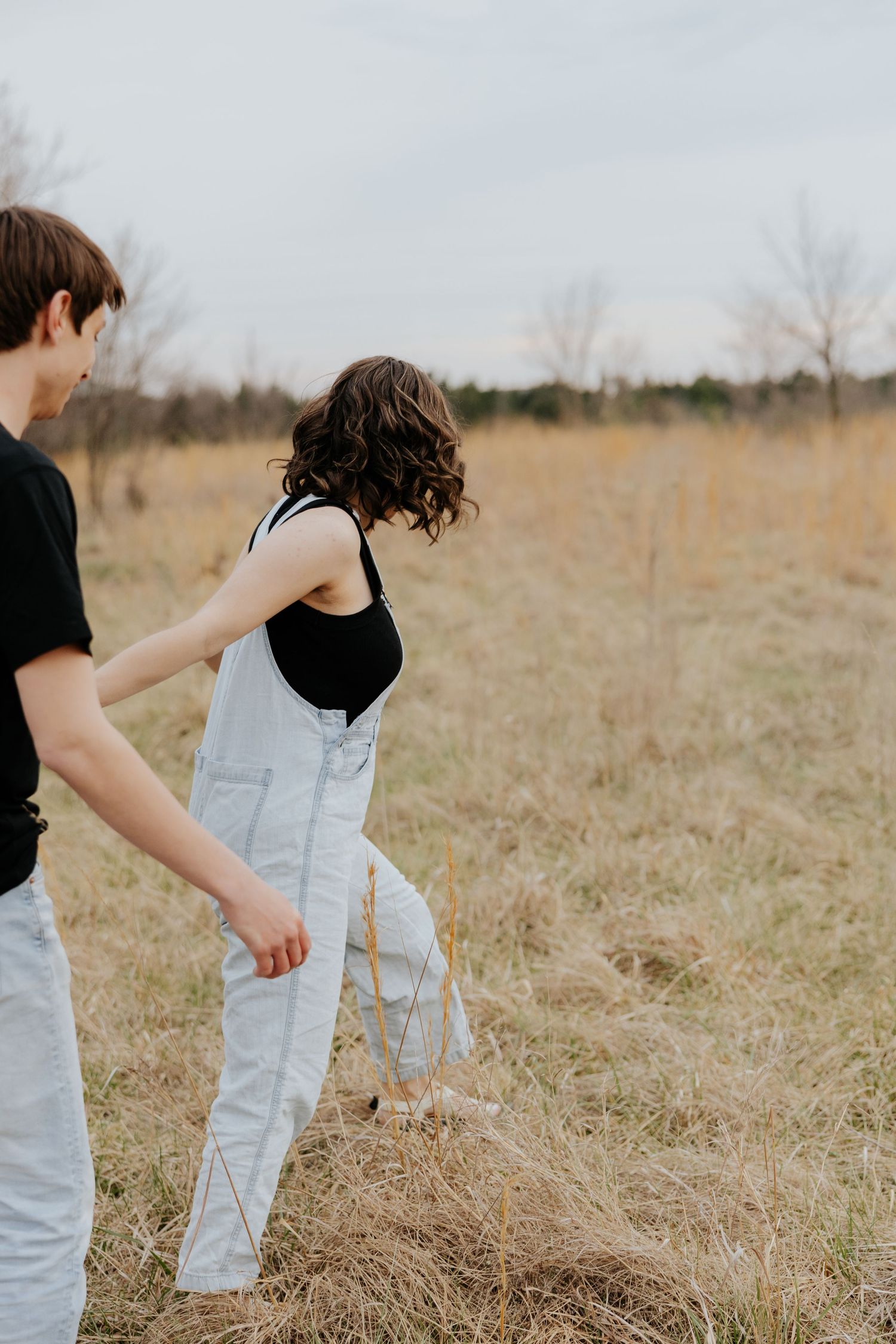 A couple walks through a golden field, woman in white overalls and black top, man in black shirt, overcast sky.