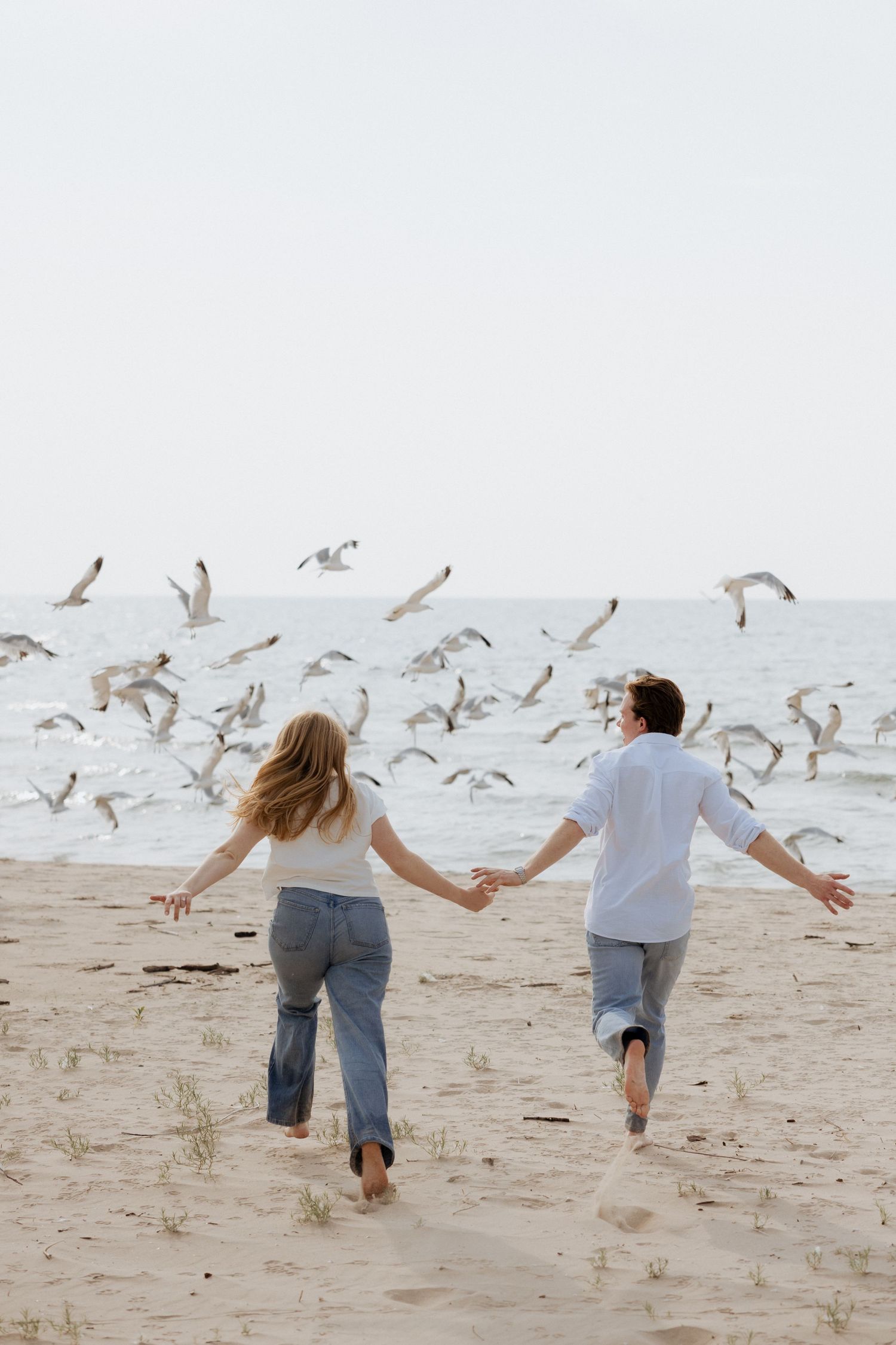 Couple holding hands running barefoot on sandy beach as a flock of seagulls takes flight around them.