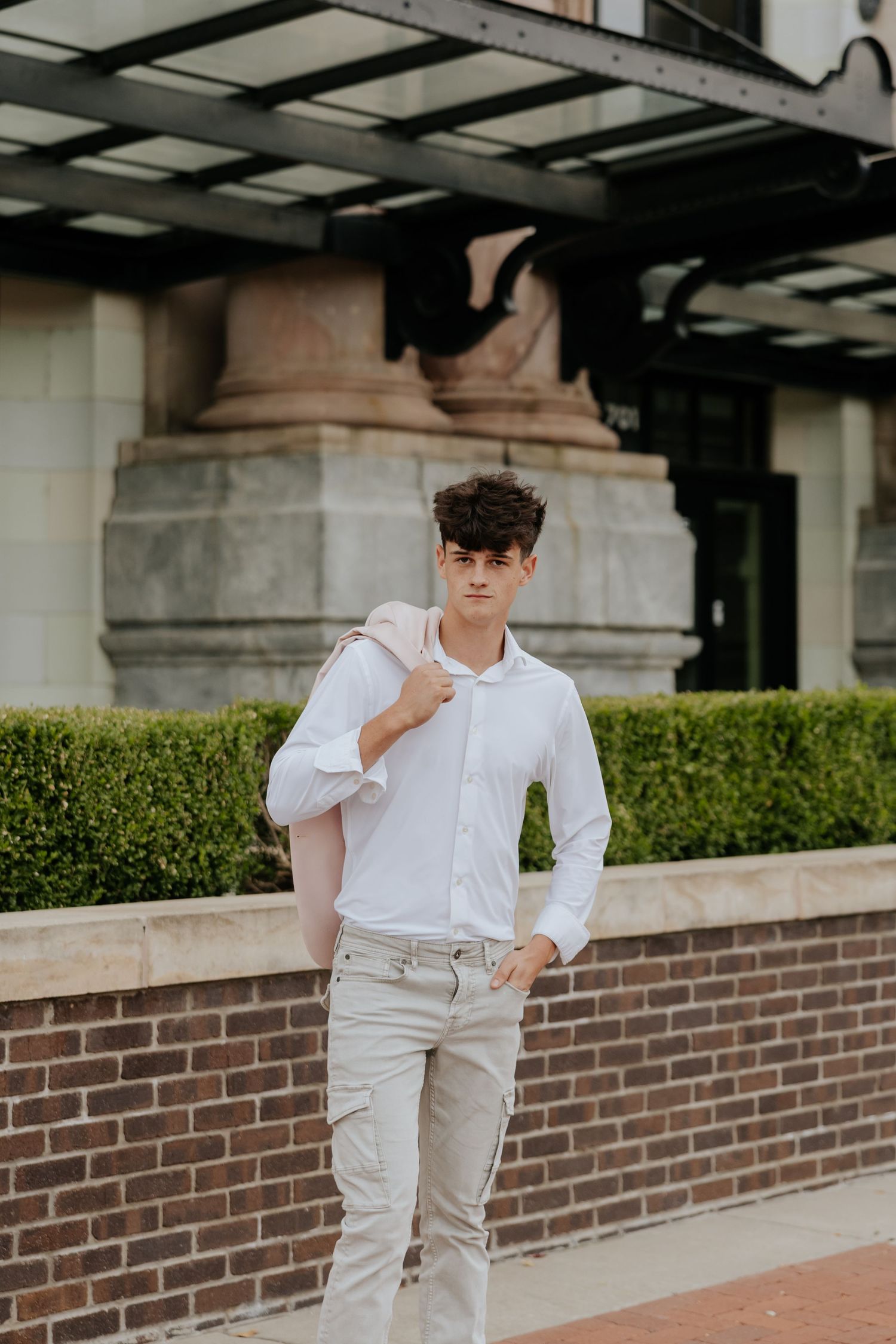Young man in white shirt carries pink blazer over shoulder while posing near brick wall and hedges.