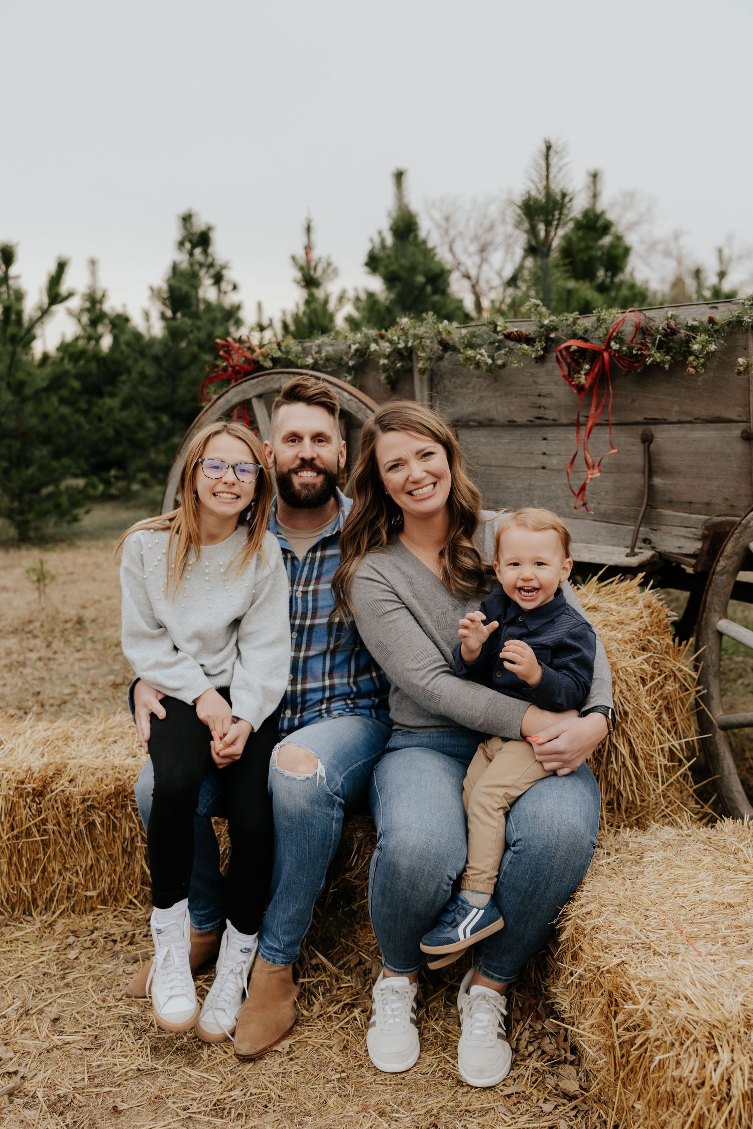 Family of four sitting on hay bale at Christmas tree farm with festive wagon.