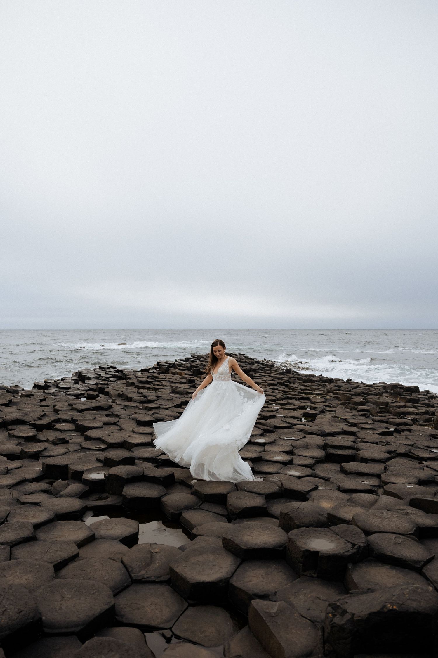 Bride in billowing white wedding dress walking on hexagonal basalt rock formations by the sea.