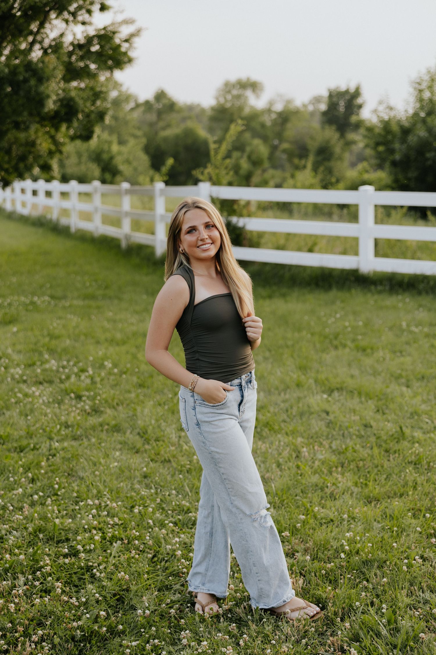 Smiling teenage girl poses in dark tank top and wide-leg jeans beside a white fence in a green field.