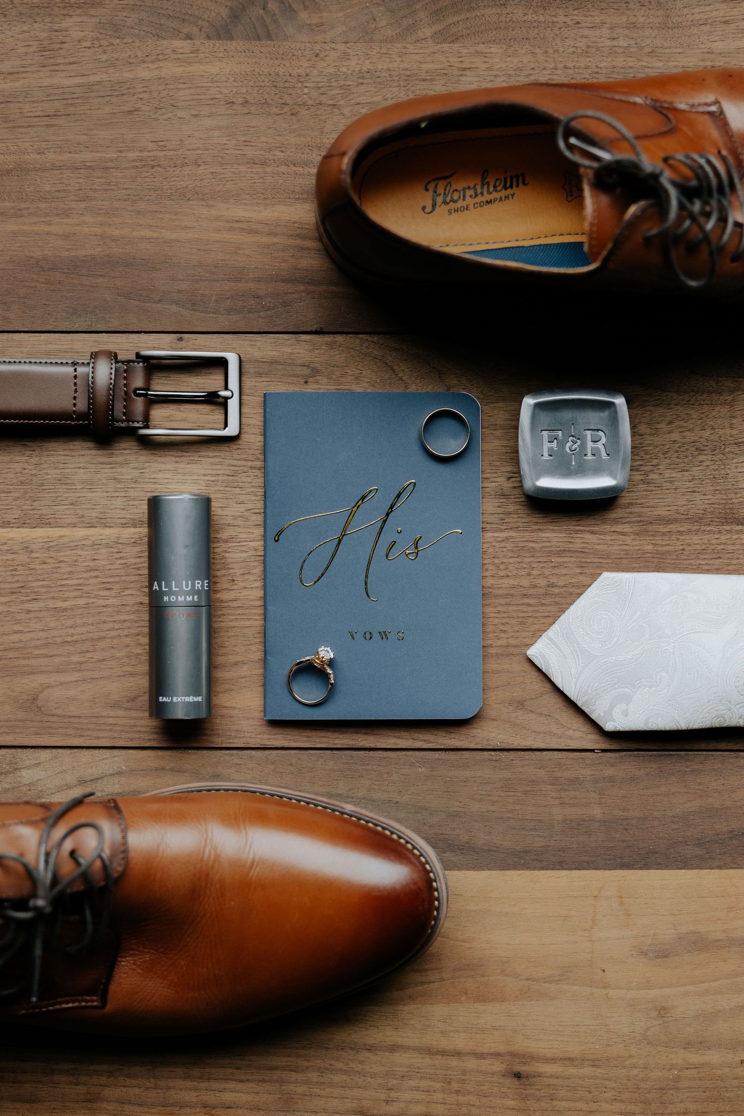 Flat lay of groom wedding accessories including brown leather shoes, belt, cologne, rings, and vow book.