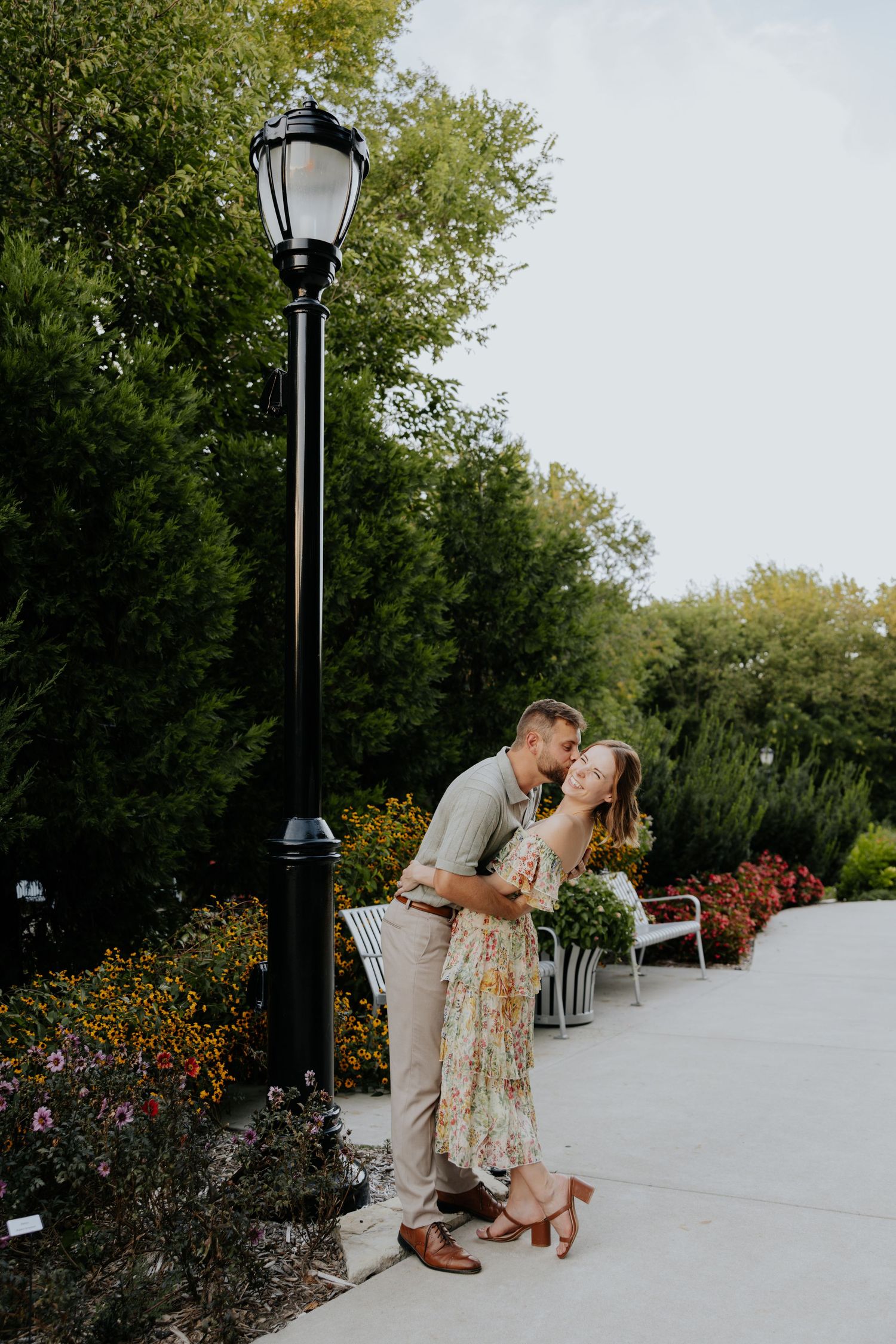 Couple shares a romantic kiss beside a vintage lamp post in a colorful garden setting.