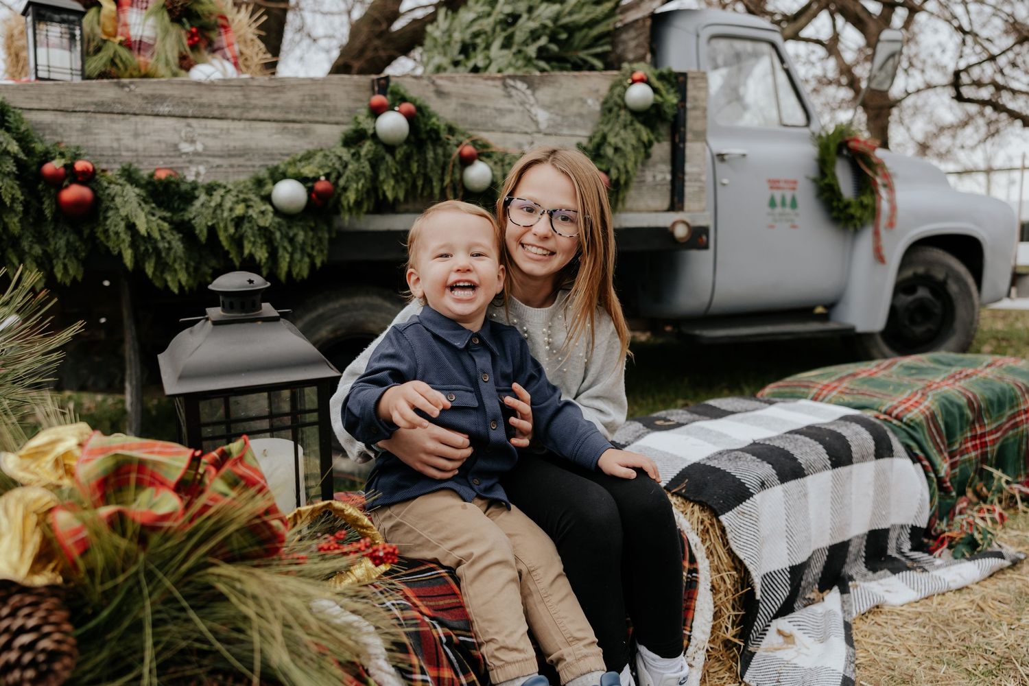 Woman with glasses holding laughing toddler at outdoor Christmas tree farm display.