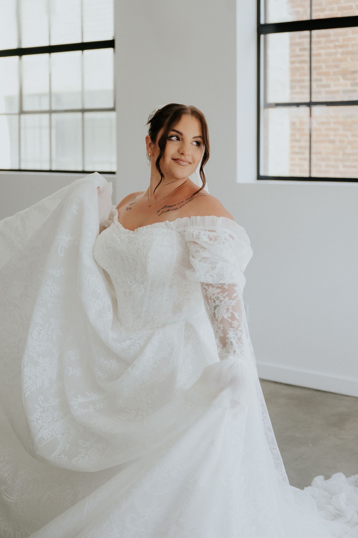 Bride in off-shoulder lace wedding gown twirling dress train in bright windowed room.