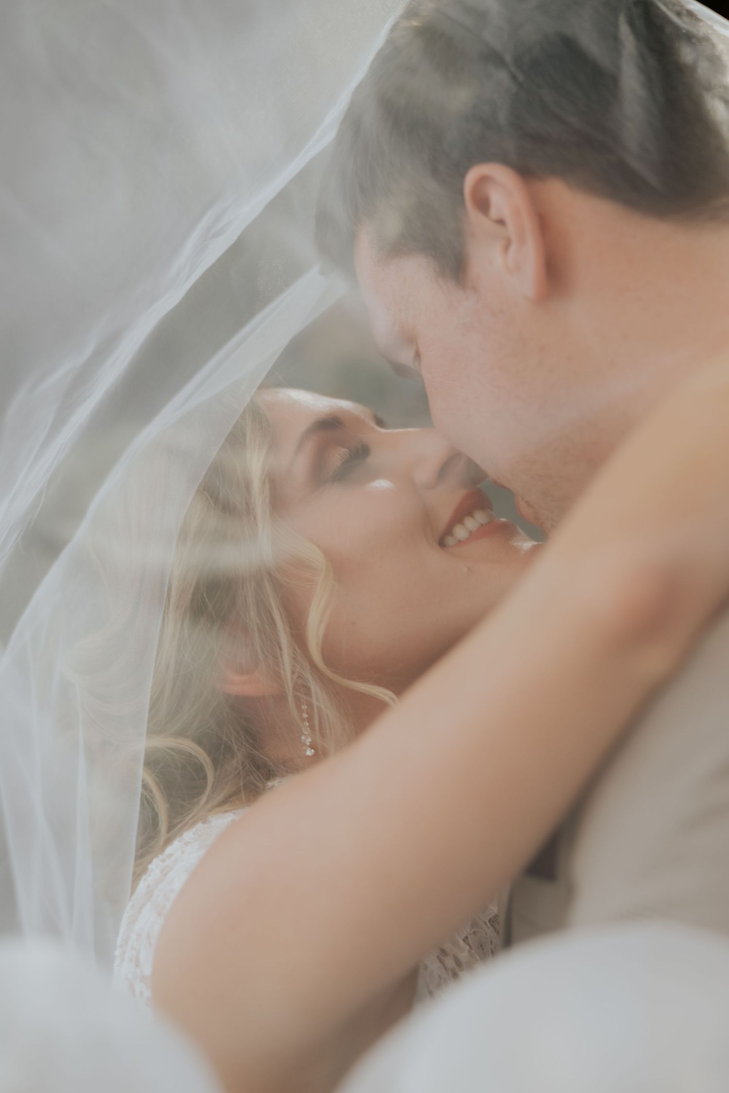 Smiling bride gazes up at groom while wrapped together under a soft white wedding veil.