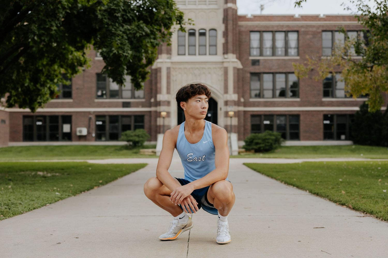 Teen boy crouches on a school pathway in athletic wear with a brick school building behind him.