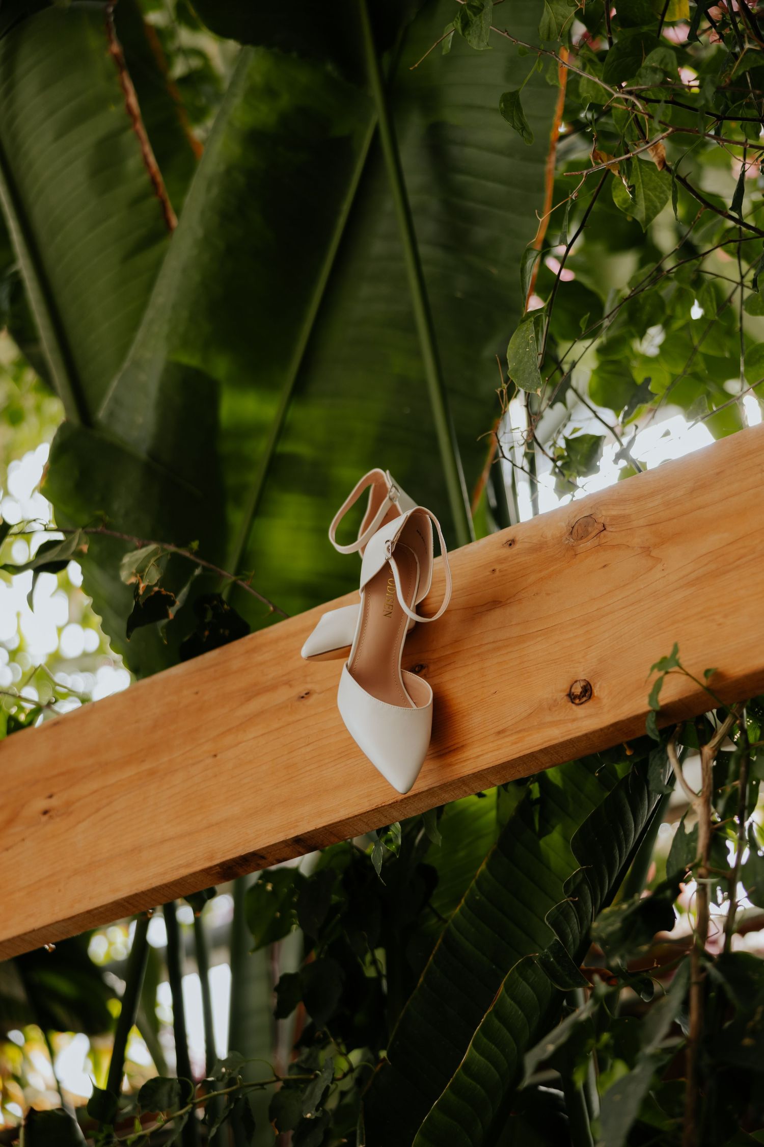 White bridal heels with ankle straps resting on wooden beam surrounded by tropical green leaves.