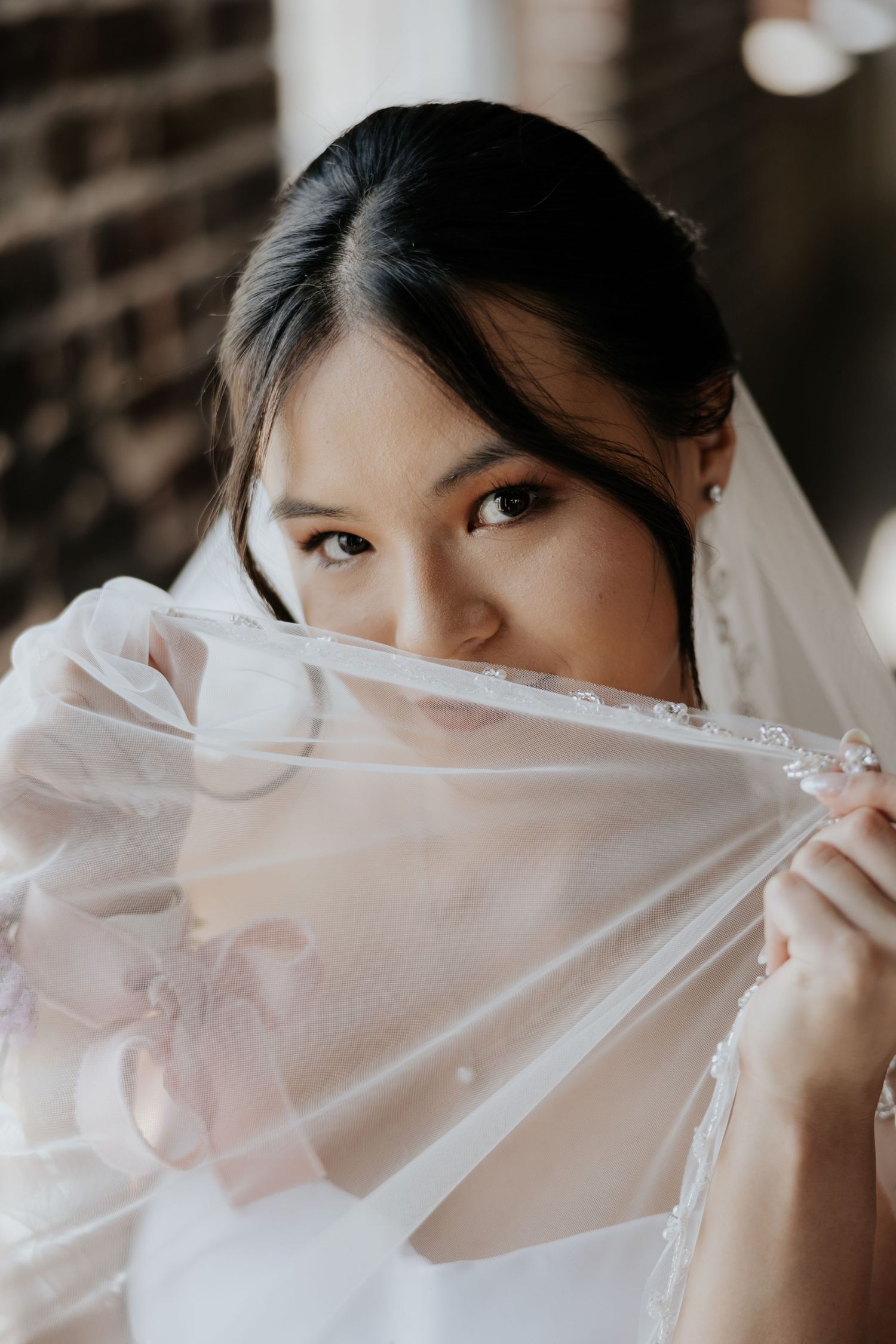 Asian bride with embroidered veil covering face, gazing at camera with soft expression.