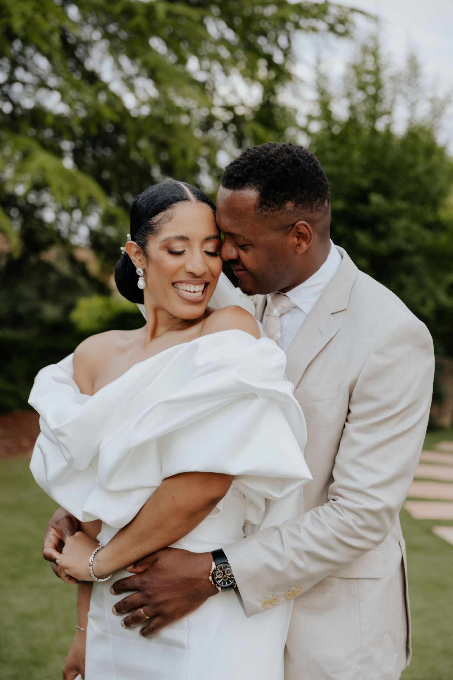 Bride in off-shoulder white gown embraces groom in beige suit outdoors at wedding.