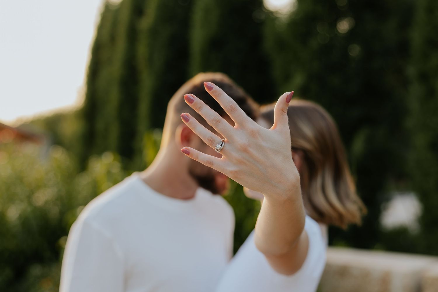 Woman proudly displays diamond engagement ring on her hand while couple kisses in soft sunlight.