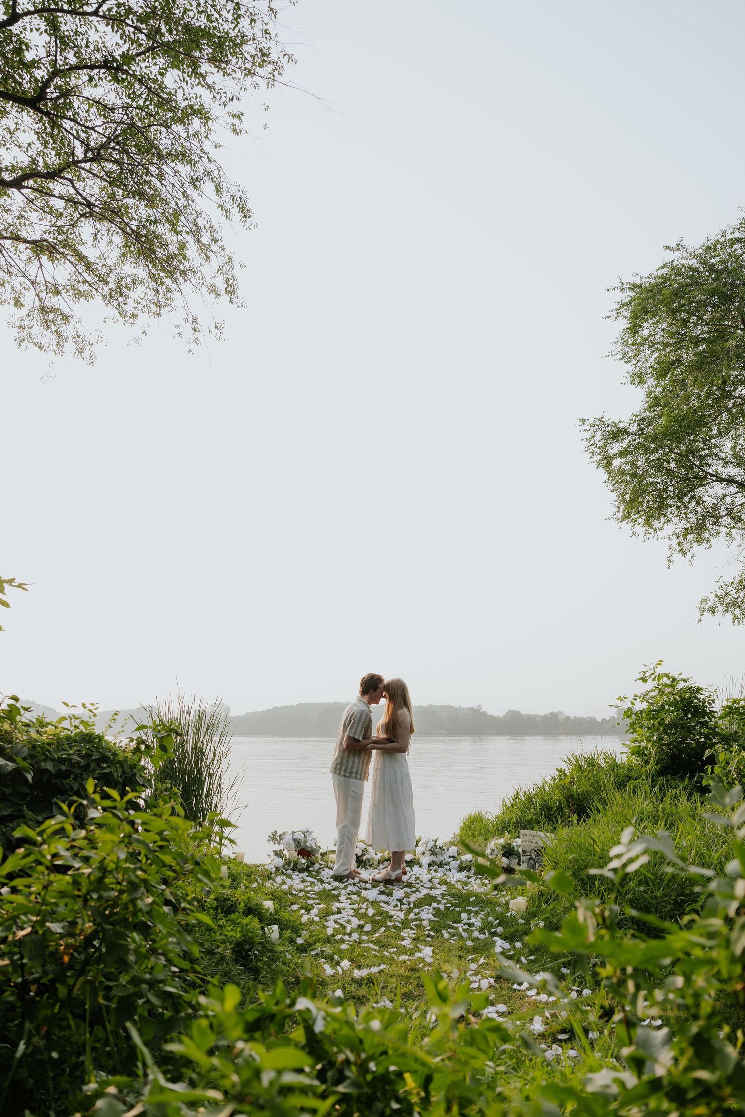 Couple kissing by a peaceful lake surrounded by lush green trees and shrubs.