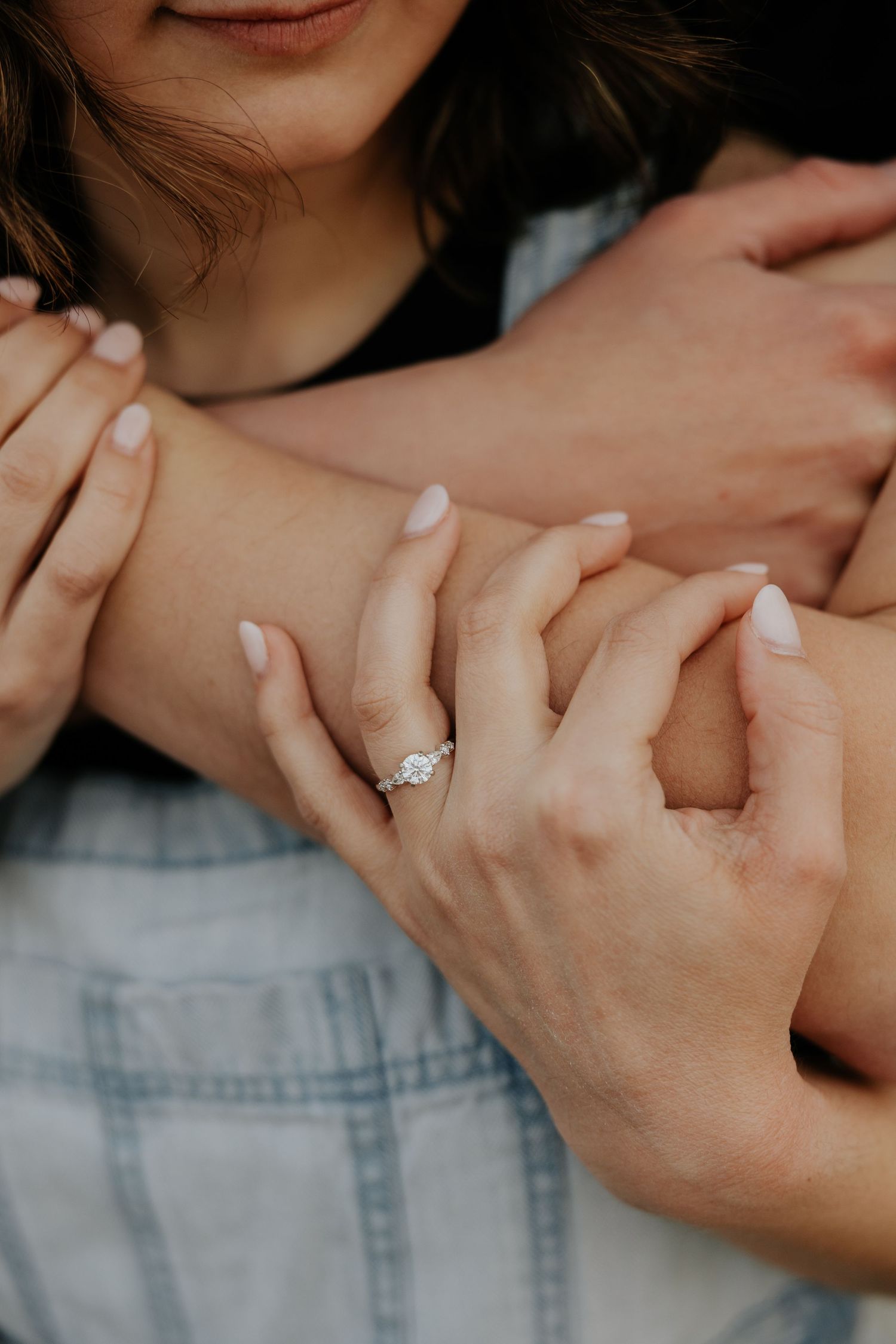 Close-up of a woman's engagement ring with diamond solitaire while couple embraces during outdoor photo session.