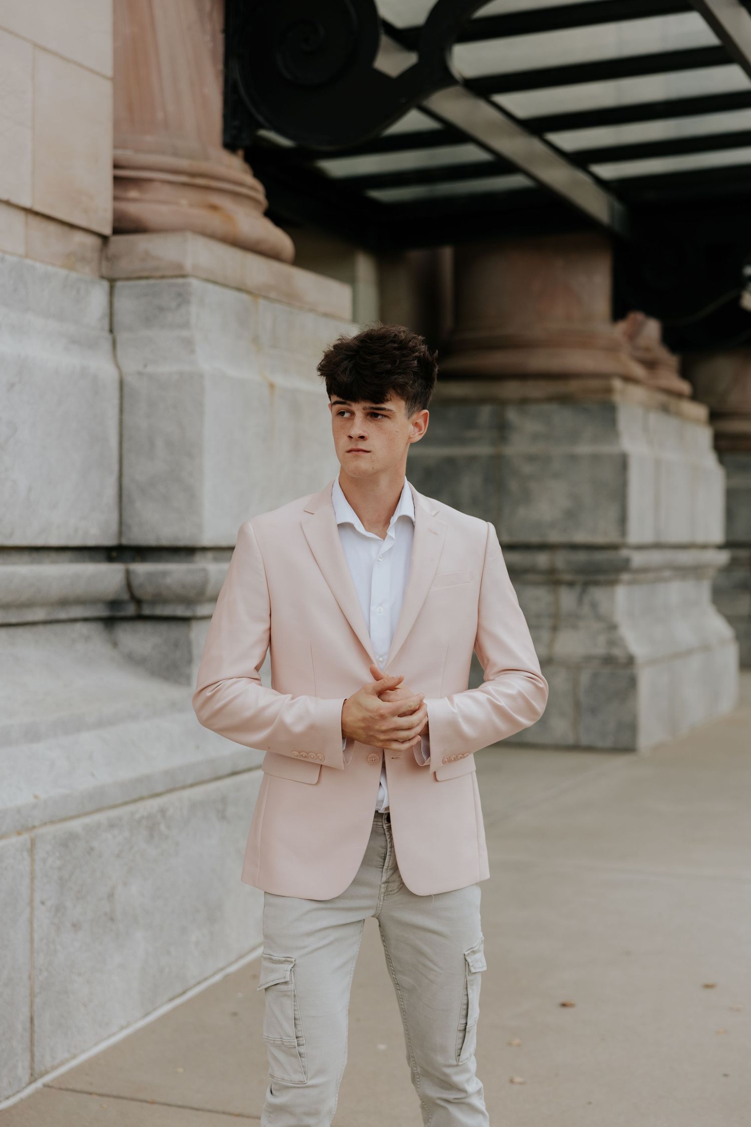 Stylish young man in pink blazer and gray pants poses near stone columns in urban setting.