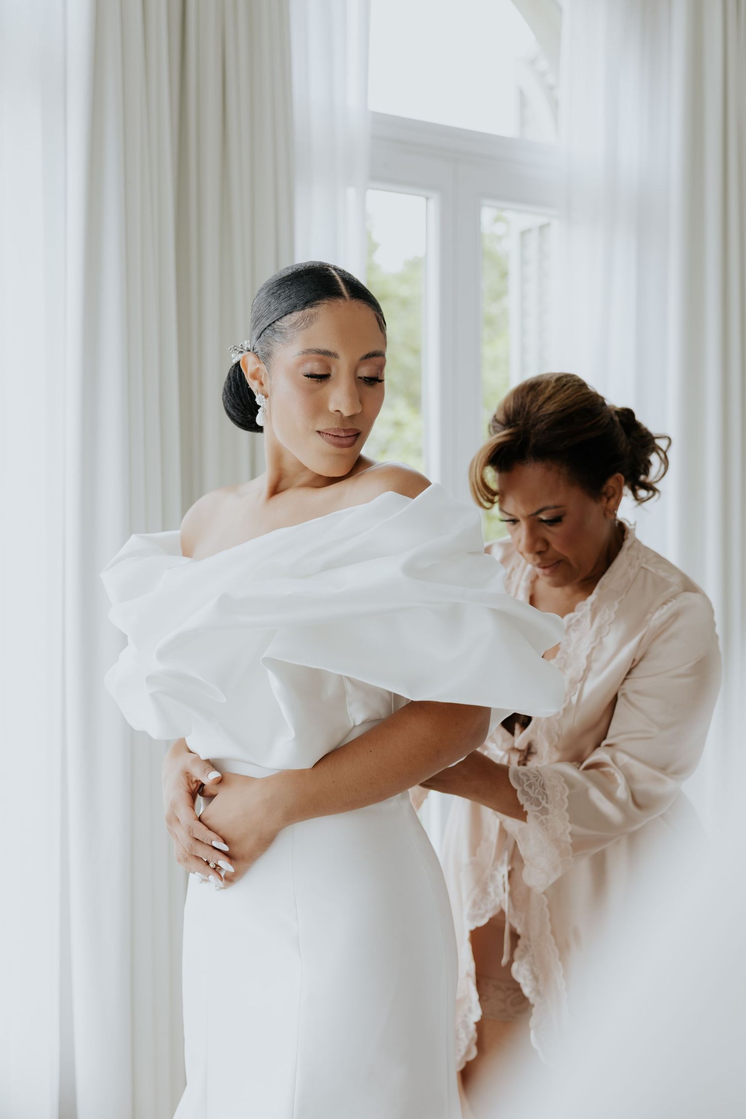 Bride in white off-shoulder gown being helped by assistant near bright window on wedding day.