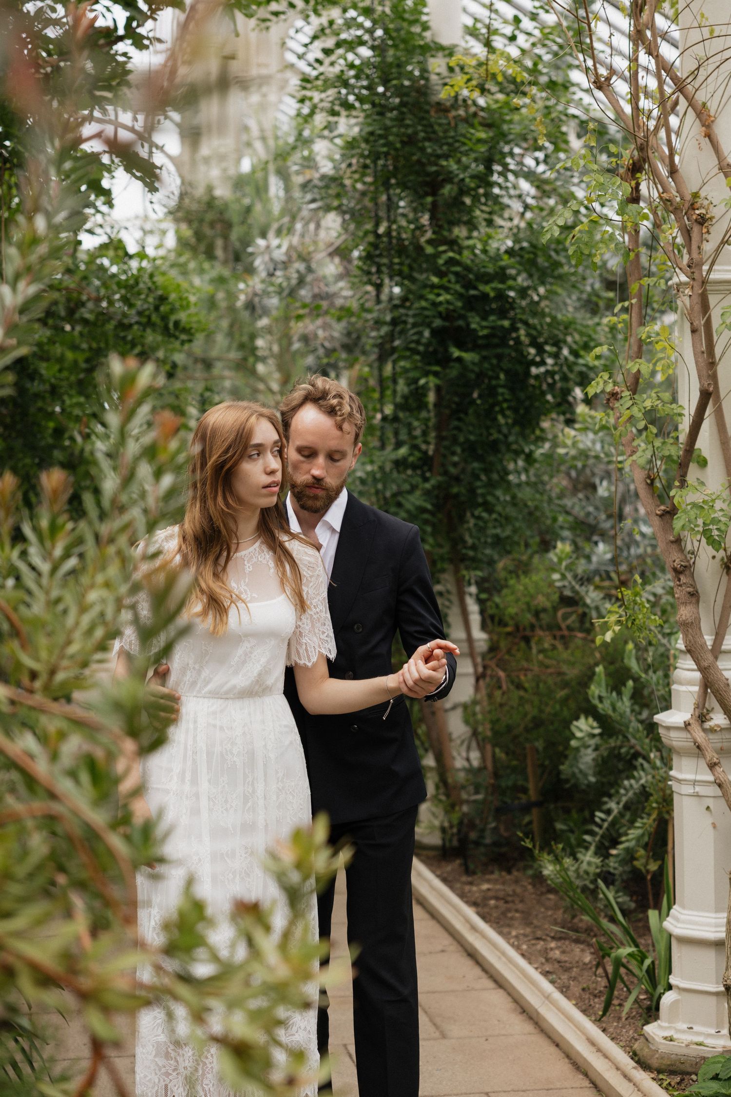 Bride and groom walking together through a lush botanical garden pathway in wedding attire.
