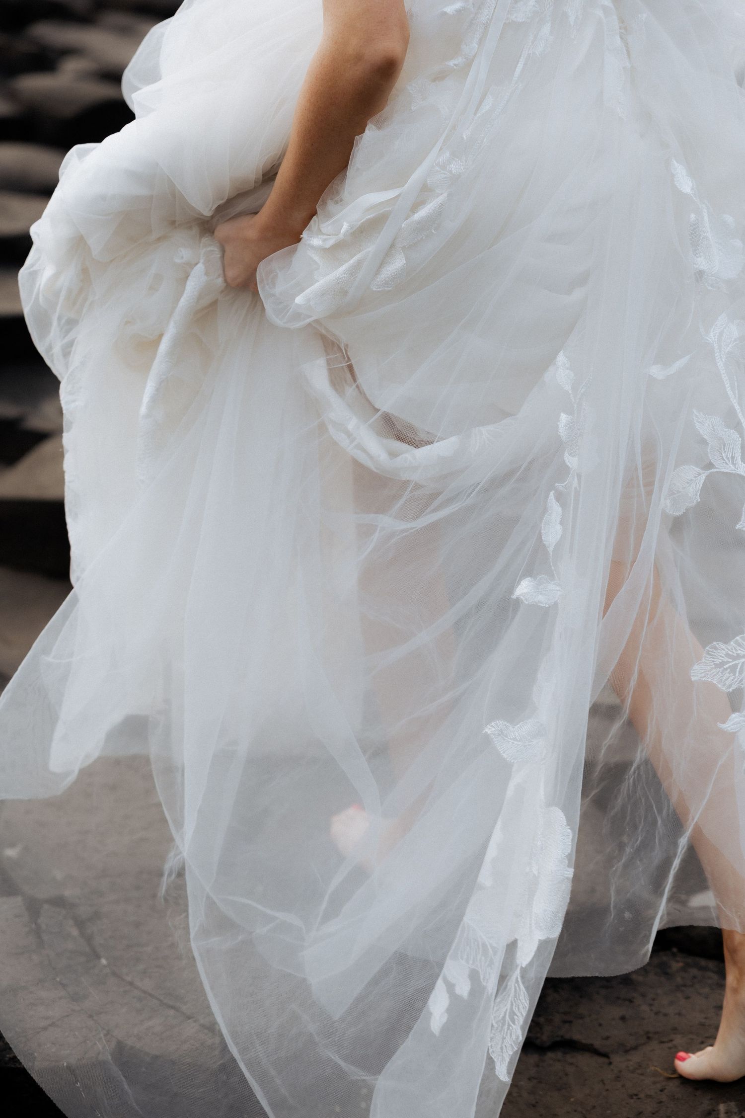 Close-up of a bride lifting flowing white tulle wedding dress skirt on rocky terrain.
