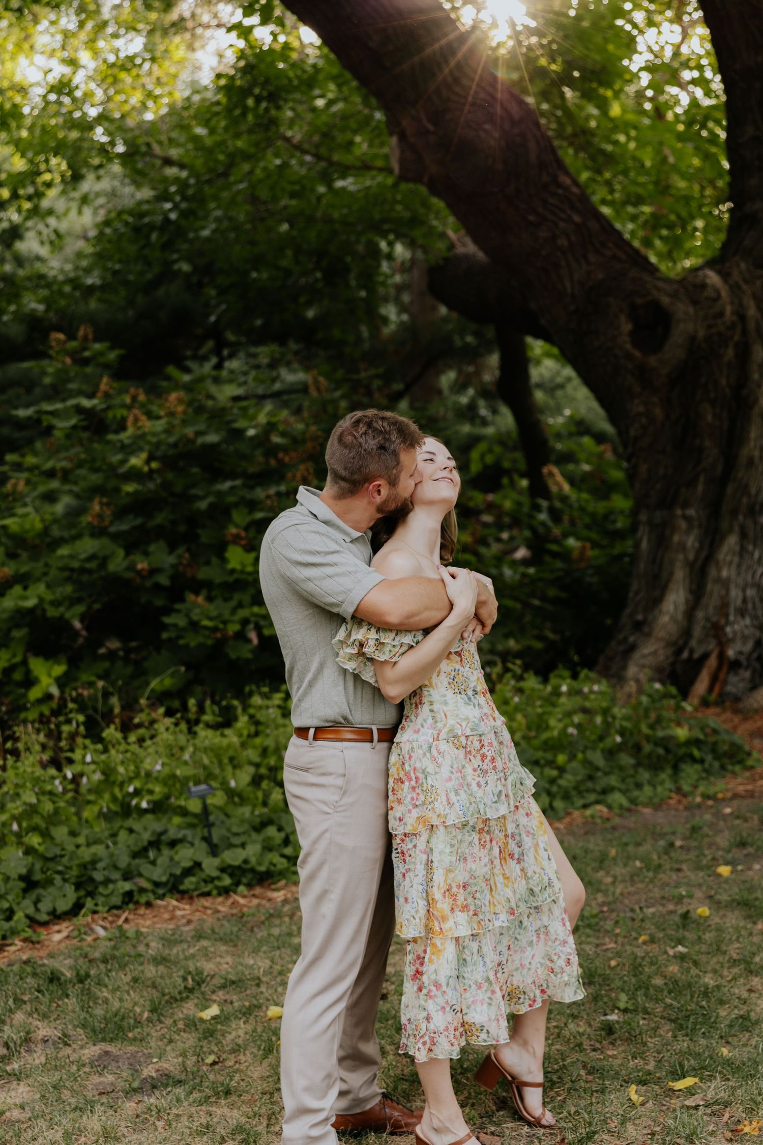 Couple embraces and kisses in golden sunlight beneath a large tree in a lush garden.