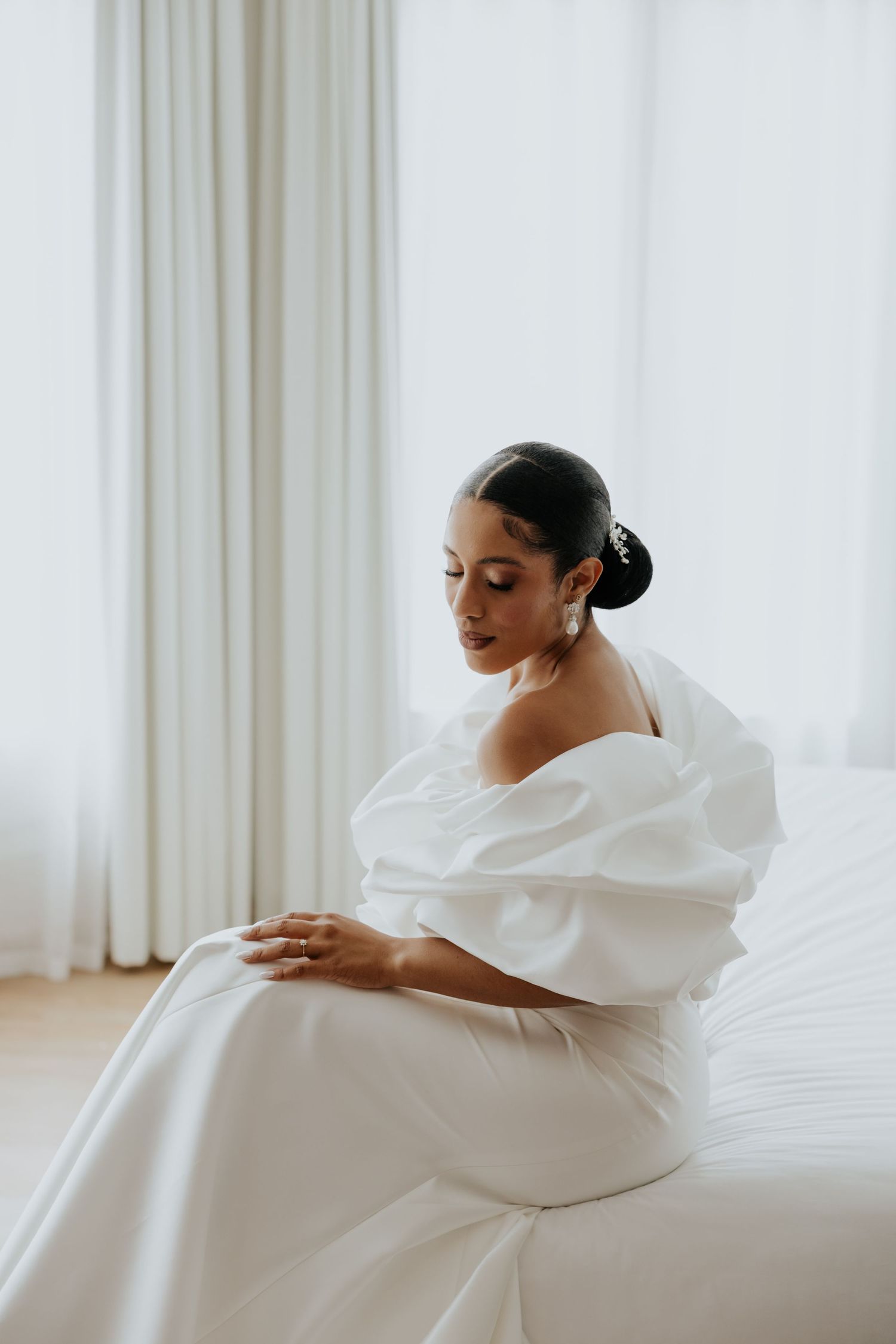 Bride in off-shoulder white gown with puff sleeves sits gracefully on bed, hair in elegant updo with pearl accessories.