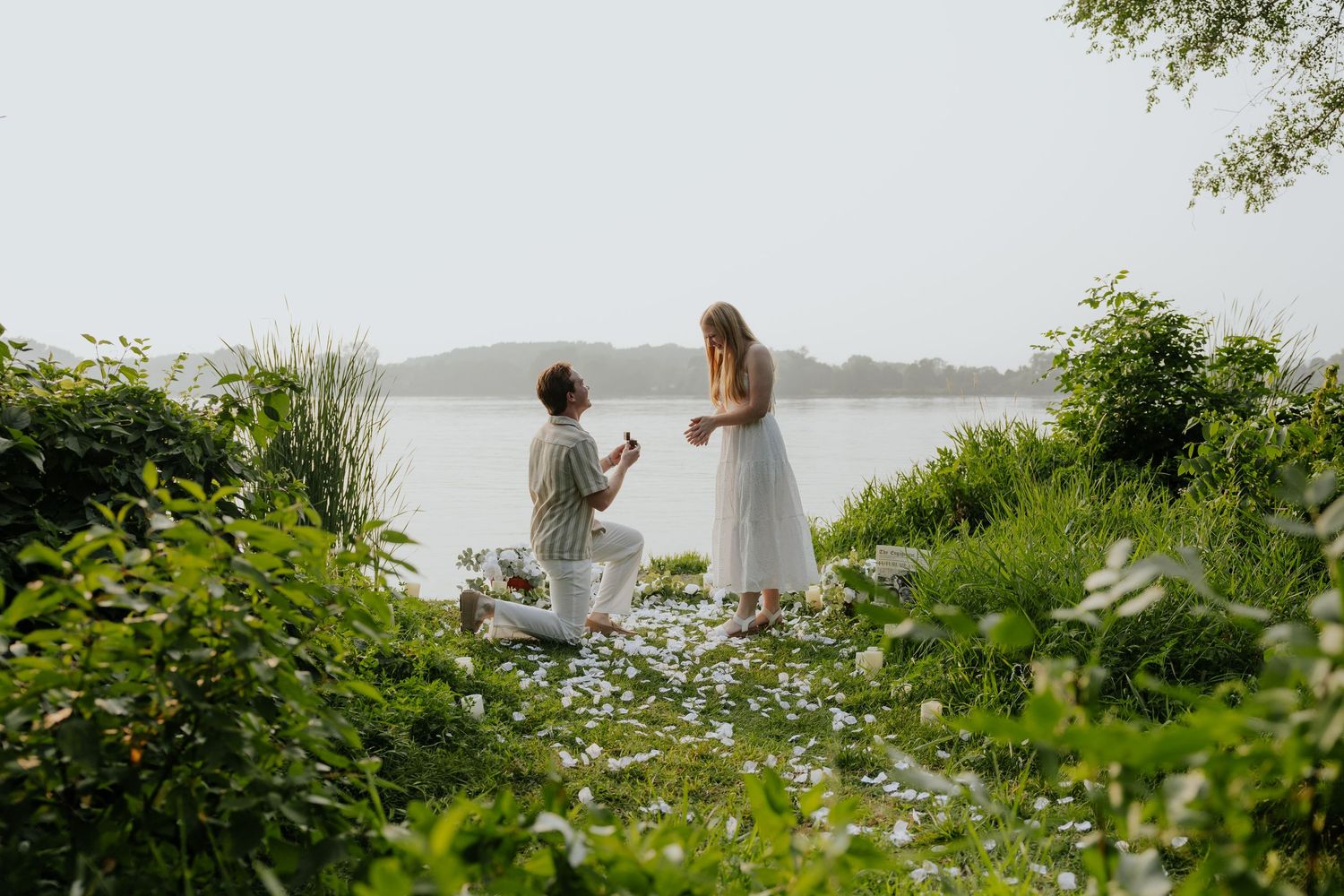 Man proposes on one knee to his partner by a scenic lakeside surrounded by wildflowers.