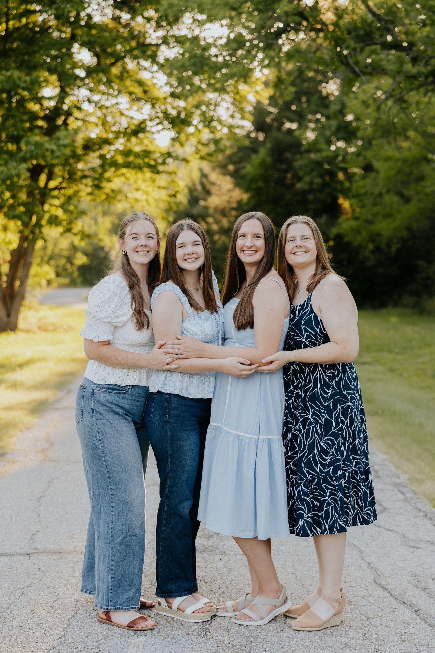 Four smiling women embrace warmly on a tree-lined path in warm golden outdoor lighting.