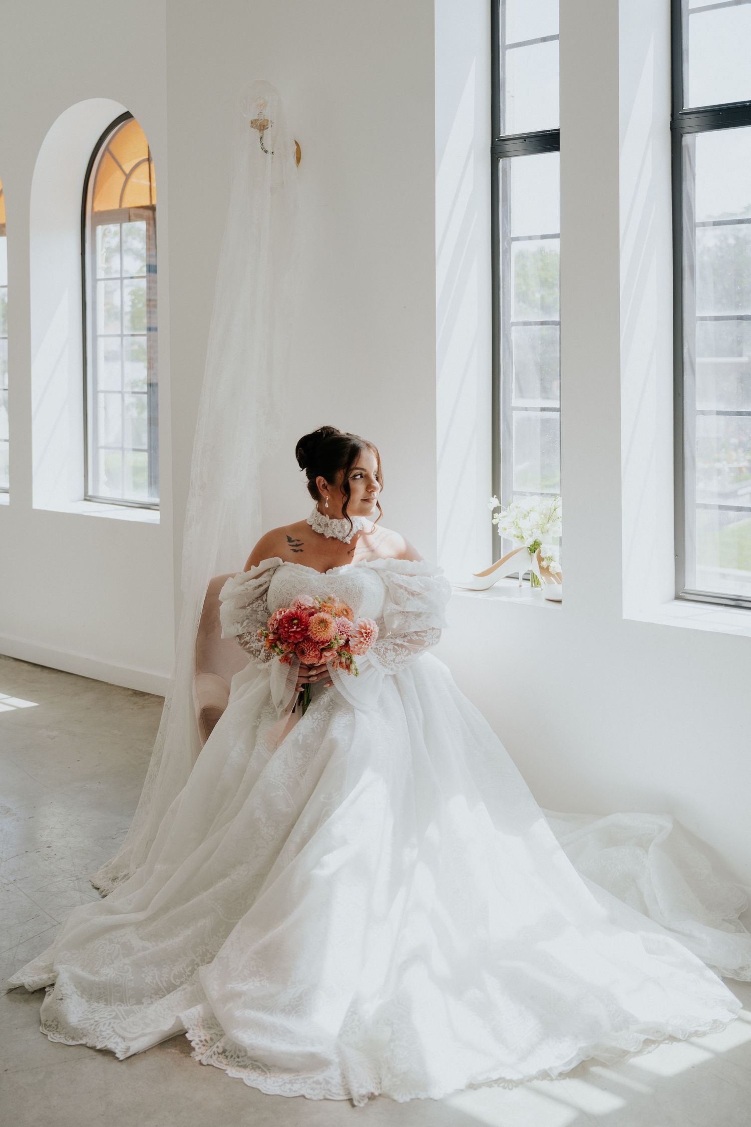 Bride in lace ball gown holding colorful bouquet seated by tall arched windows in elegant venue.
