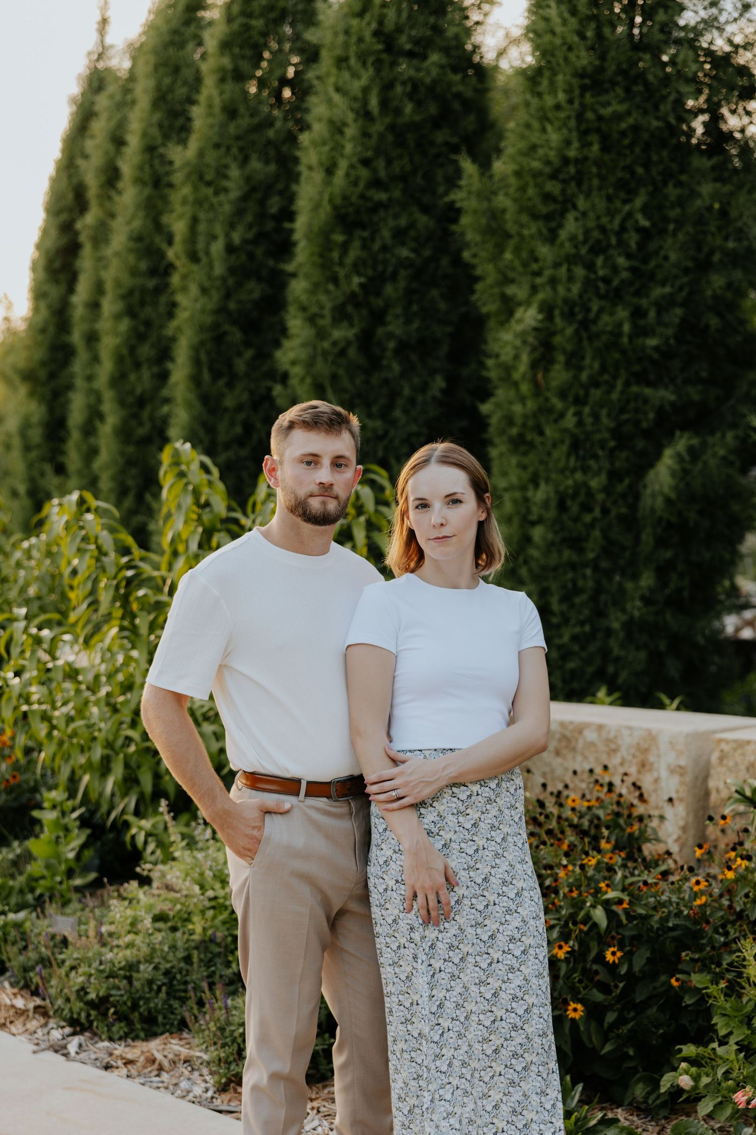 Couple stands together in white tops with serious expressions in a lush evergreen garden setting.