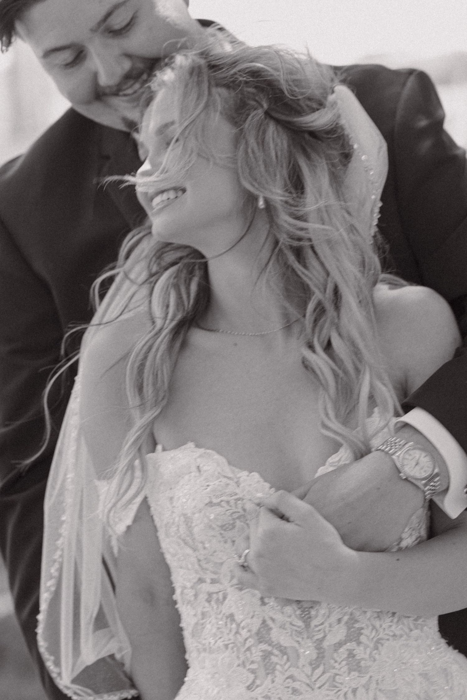 Black and white wedding portrait of a groom embracing his smiling bride in a lace gown from behind.
