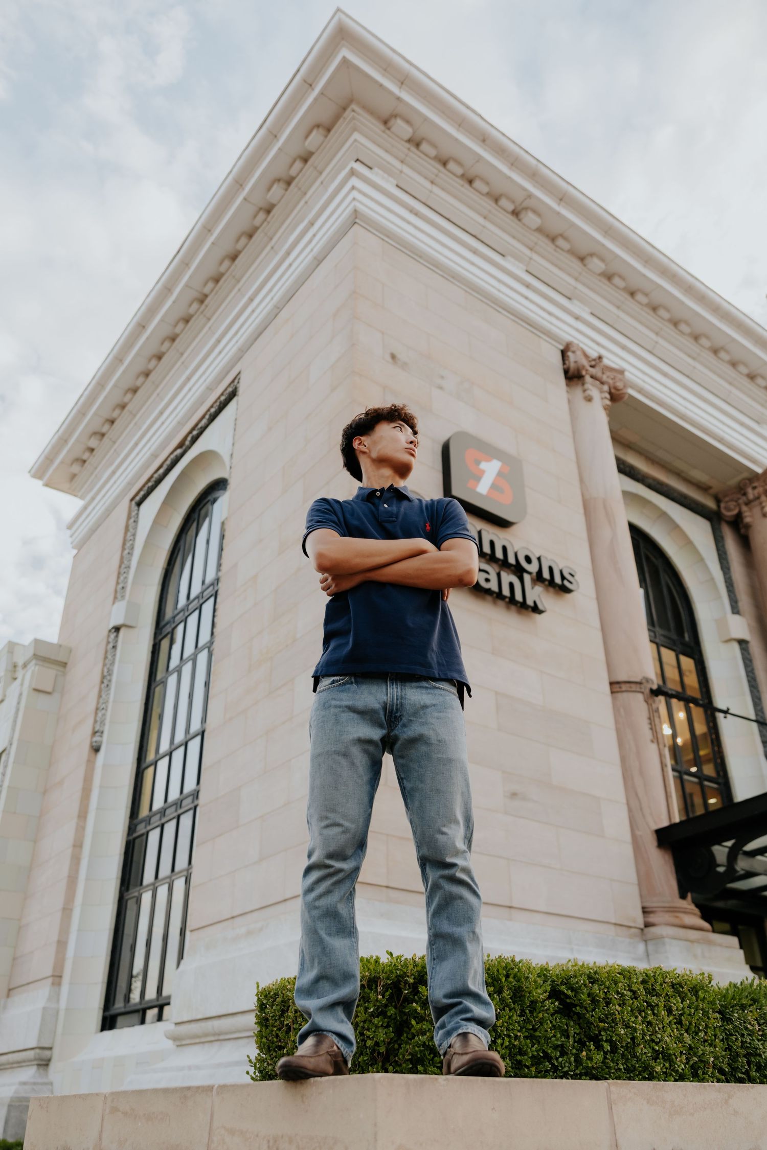 Confident teen boy stands arms crossed on steps before a classical stone bank building.