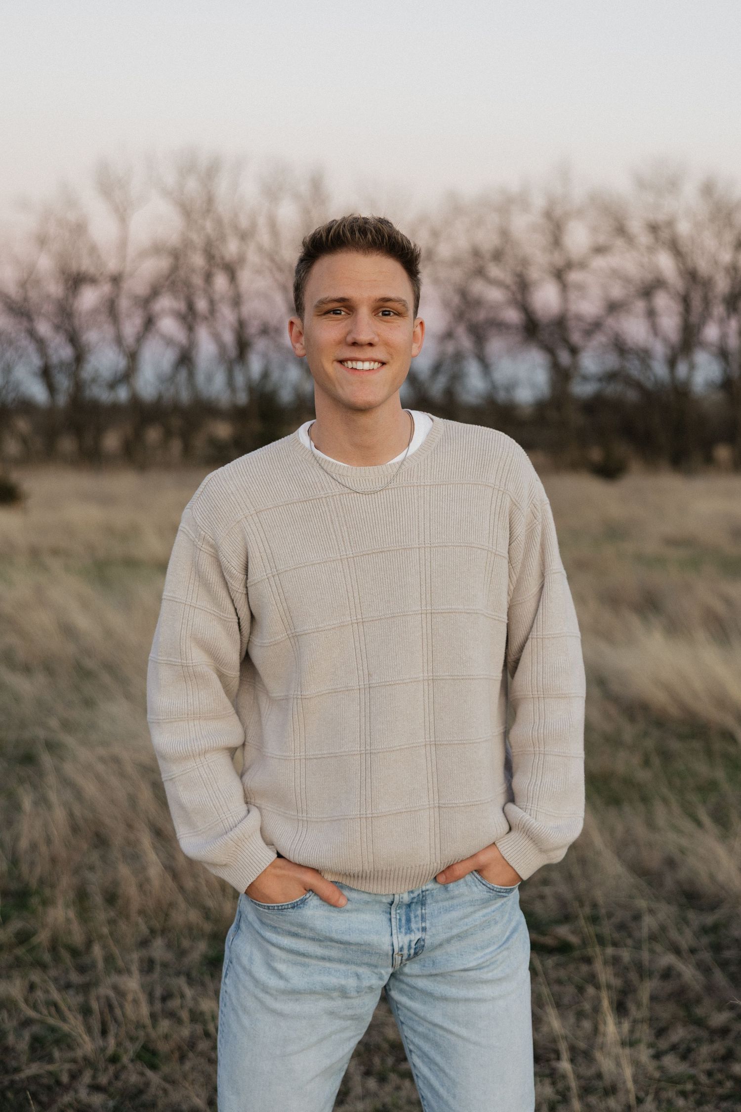 Smiling young man in beige sweater and jeans standing in an open field at golden hour.