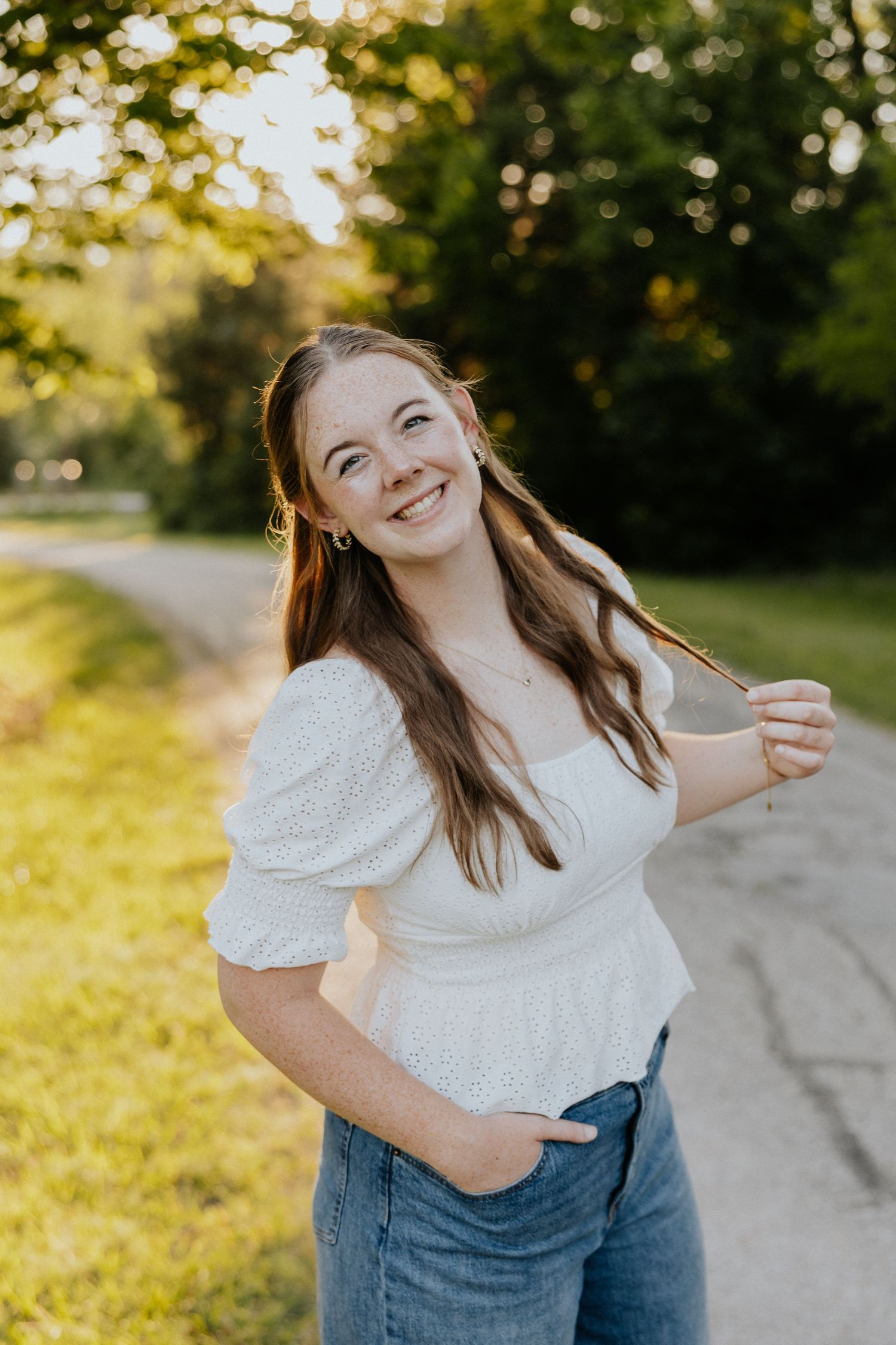 Smiling young woman in white top and jeans twirls hair on sunlit green outdoor path.