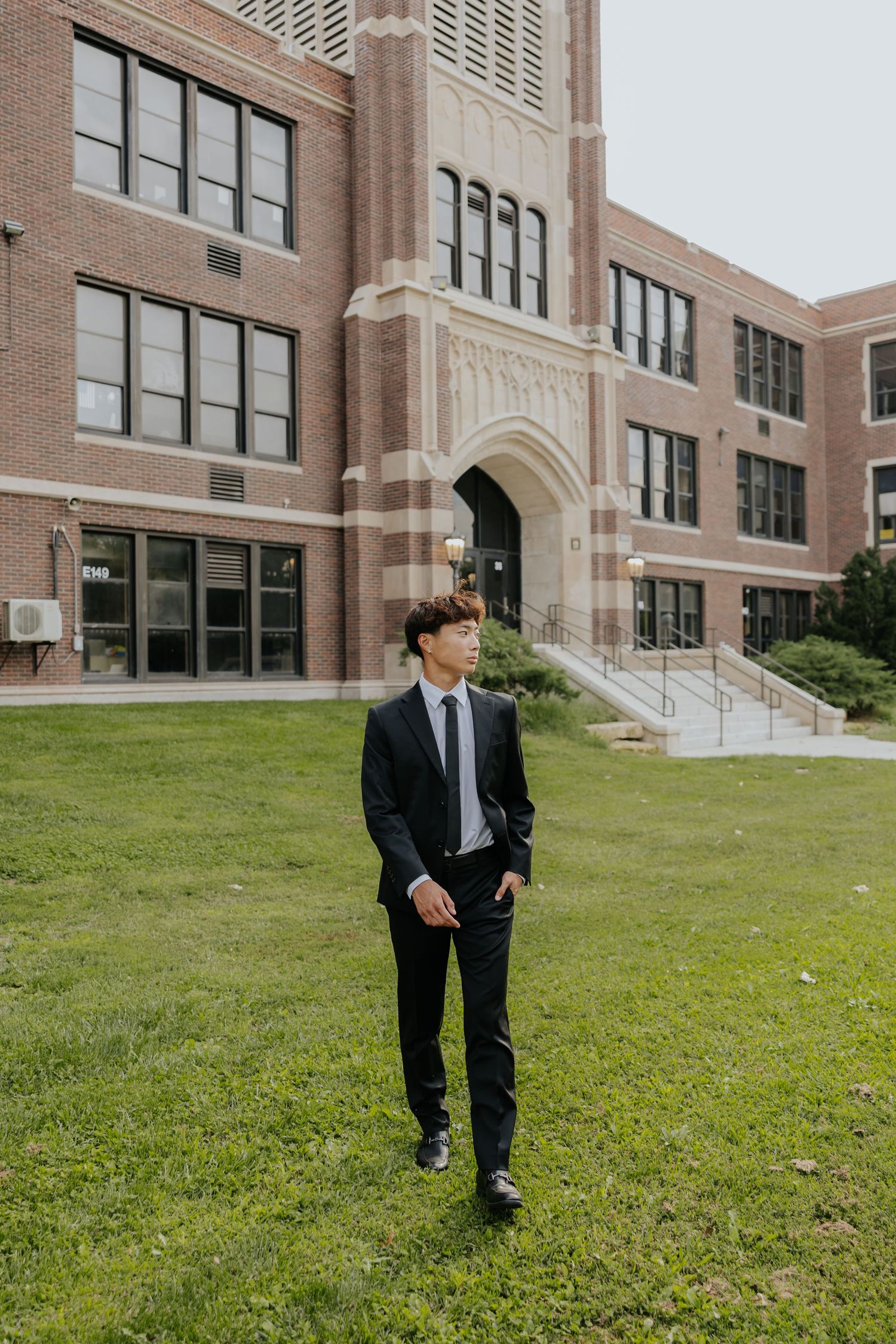 Young man in black suit standing on green lawn in front of a brick school building.
