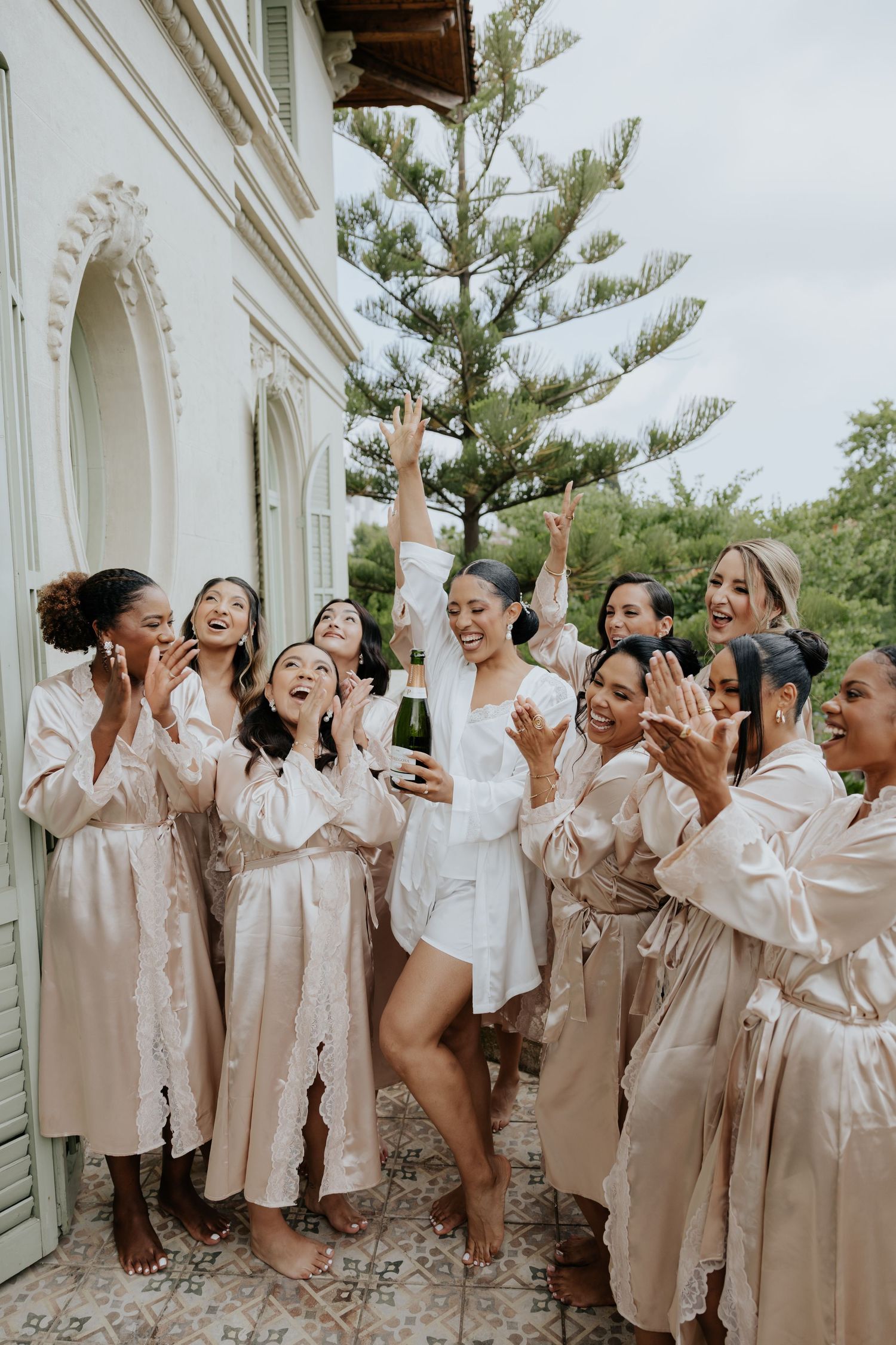 Bride in white robe celebrates with bridesmaids while popping champagne before the wedding.