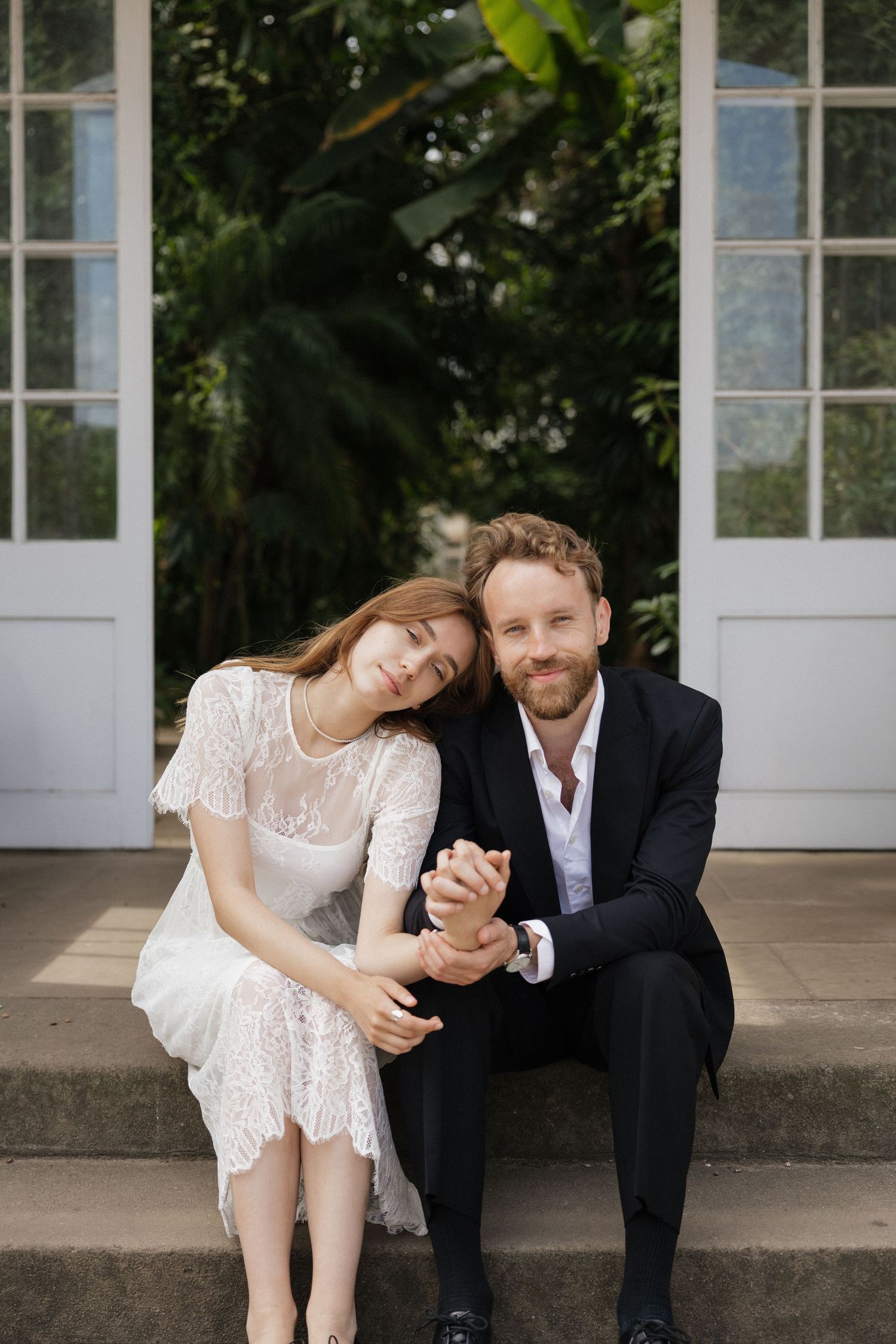 Bride resting head on grooms shoulder while holding hands seated on steps before white doors.