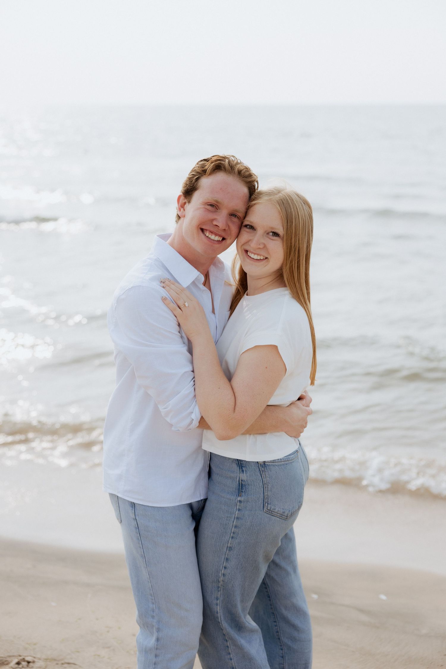 Happy couple embracing and smiling on a sandy beach with ocean waves in the background.