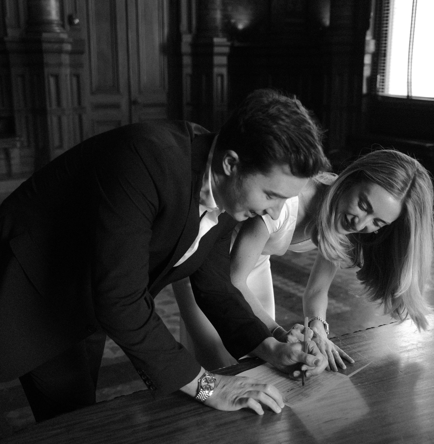 Black and white photo of bride and groom signing wedding documents at wooden table.