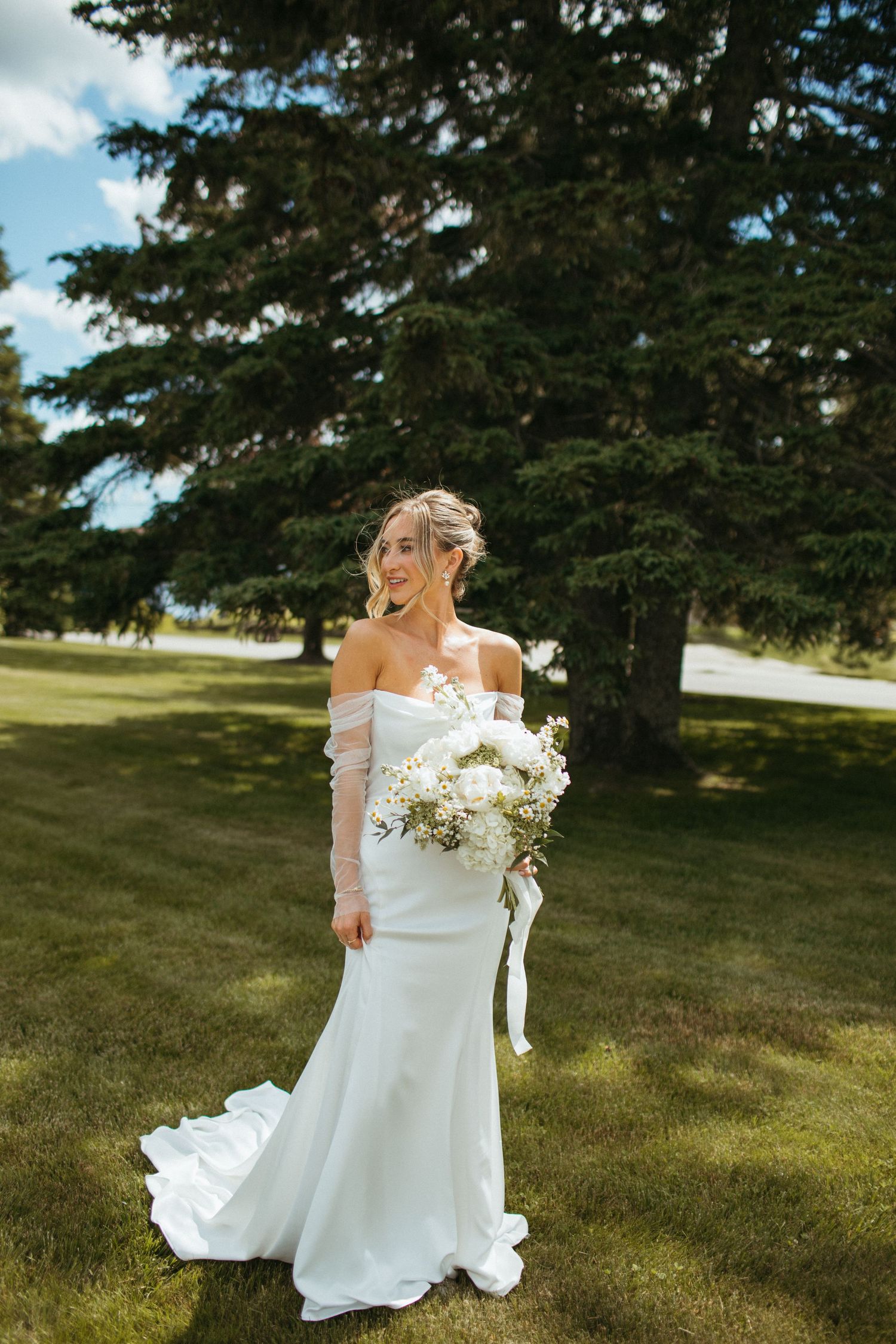 Bride in white off-shoulder mermaid gown with sheer sleeves holding white bouquet outdoors on sunny day.