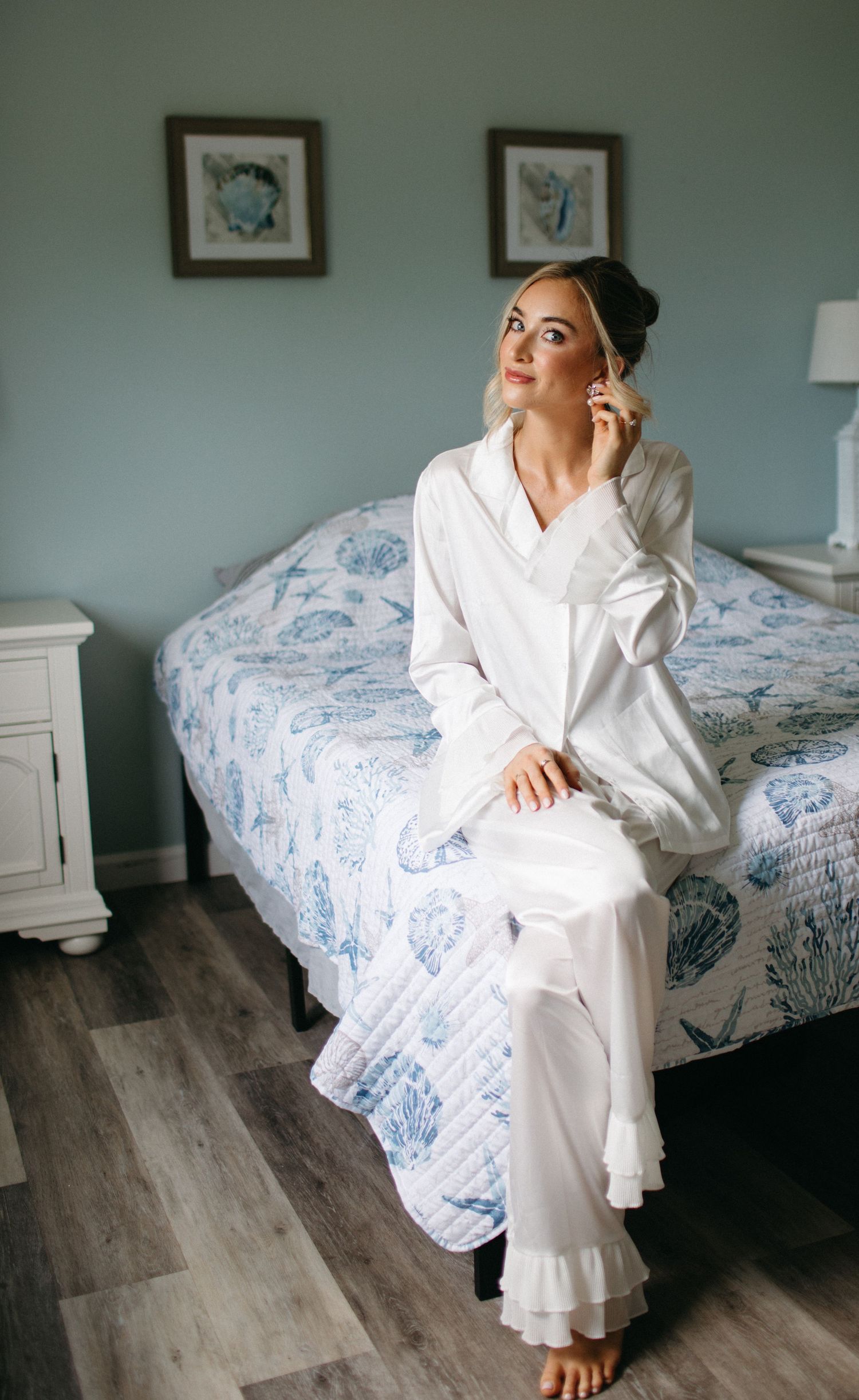 Beautiful blonde bride in white satin pajamas with ruffle hem sitting on bed getting ready.