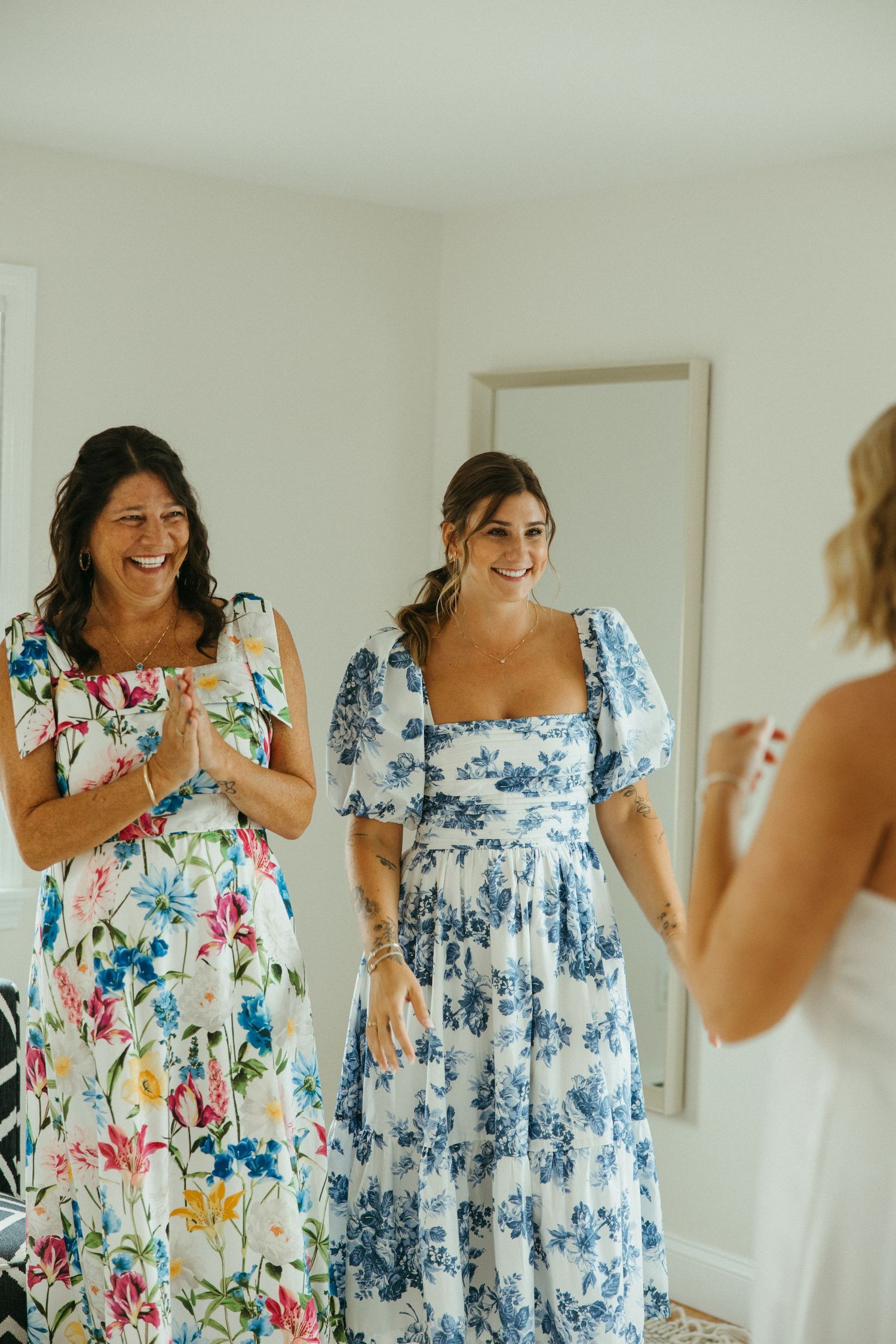 Two women in floral dresses smiling at bride during wedding morning preparations.
