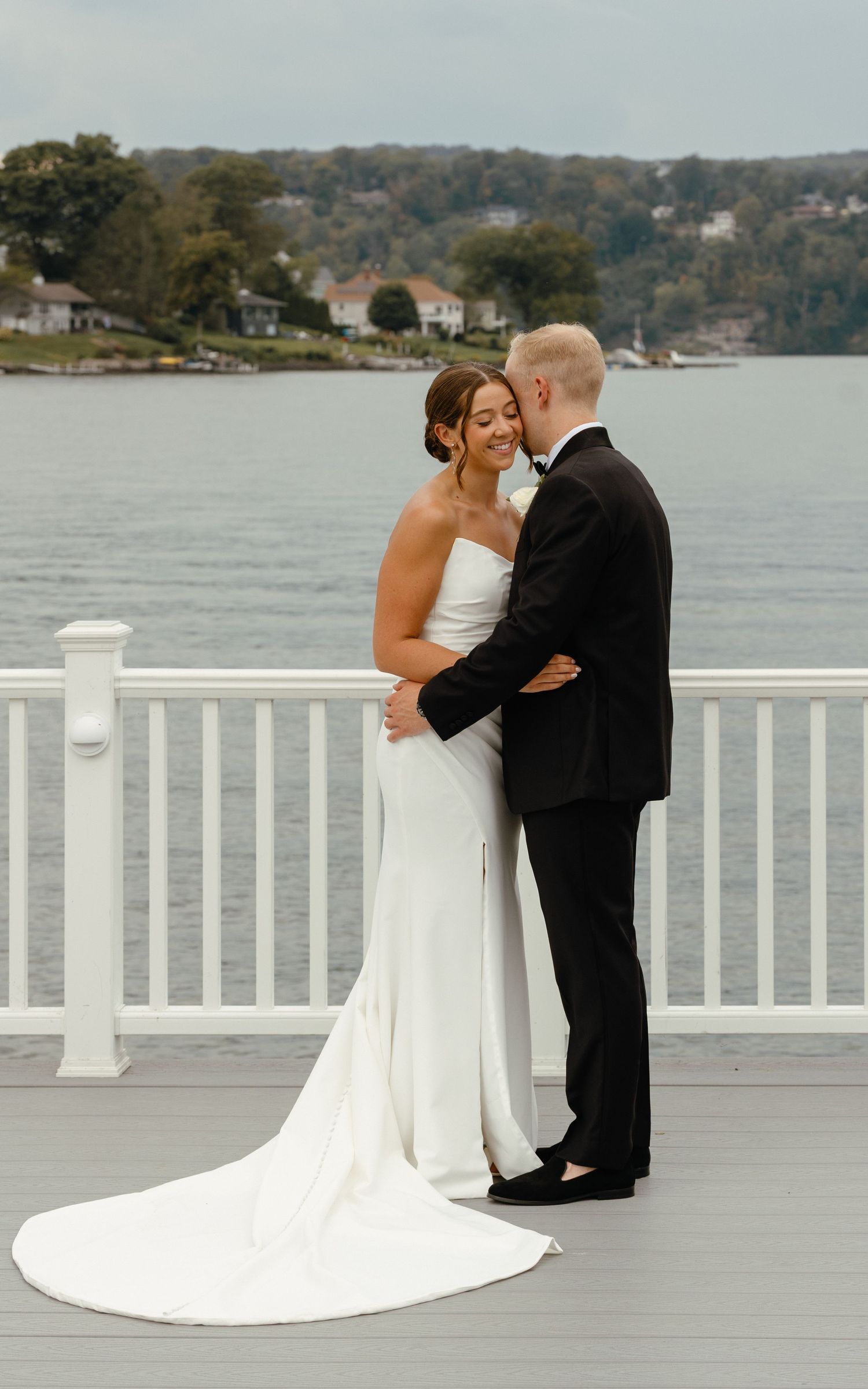 Bride and groom share a romantic moment on a waterfront deck, bride in elegant strapless gown with train.