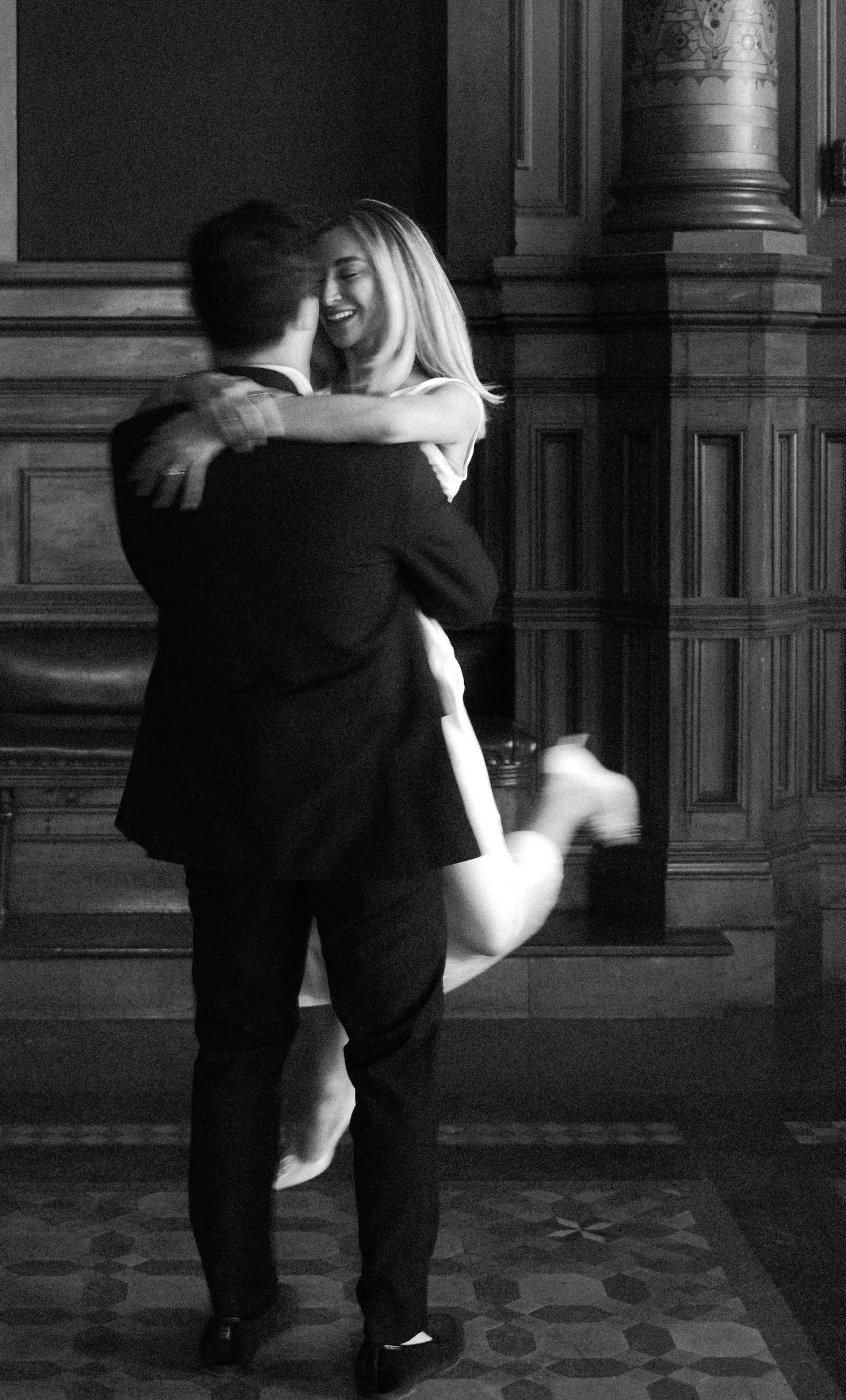 Black and white photo of a joyful couple embracing, woman laughing as man lifts her in a grand interior.film-inspired