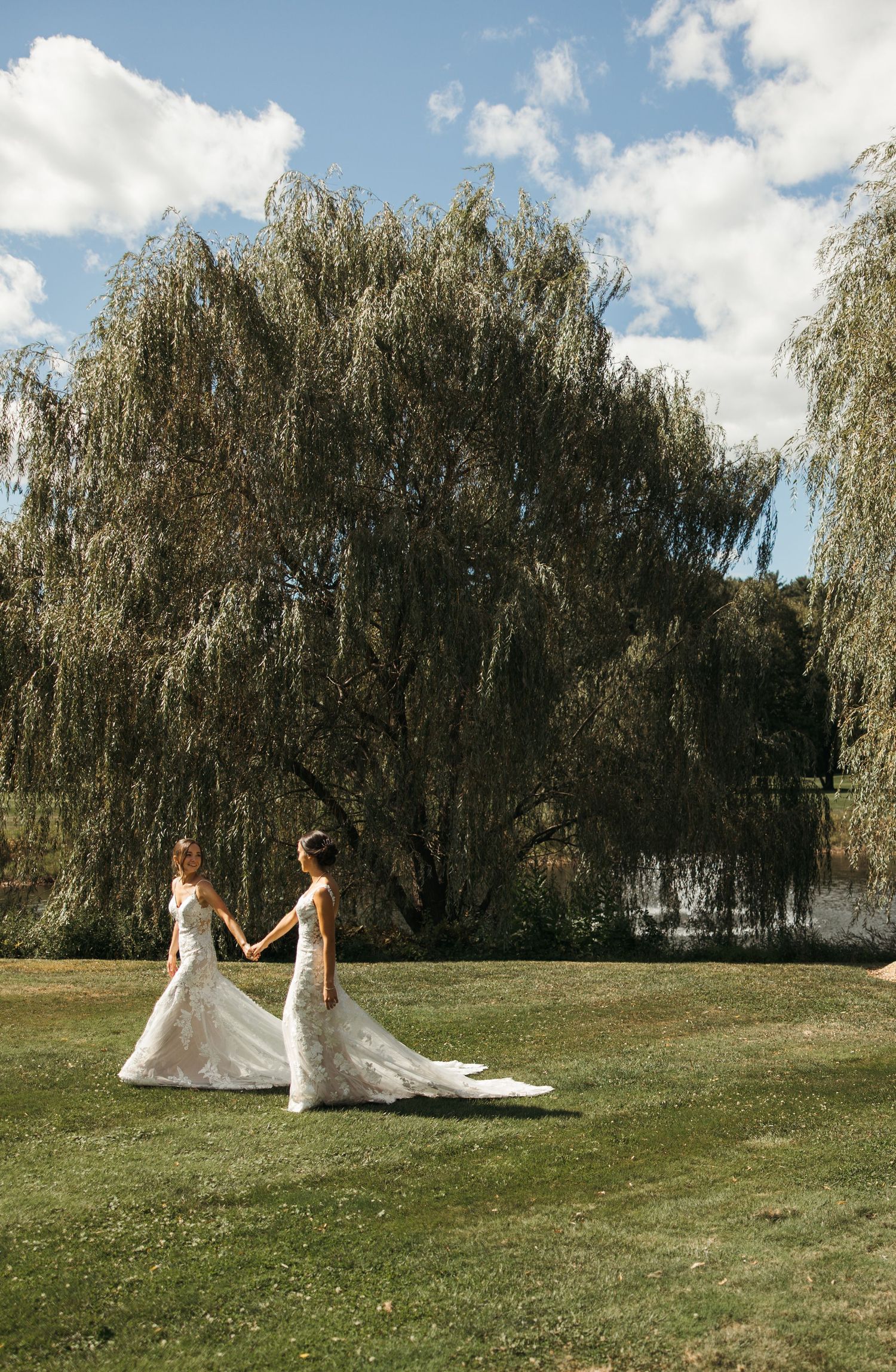 Two brides in lace wedding gowns hold hands while walking on a lawn near a large weeping willow.