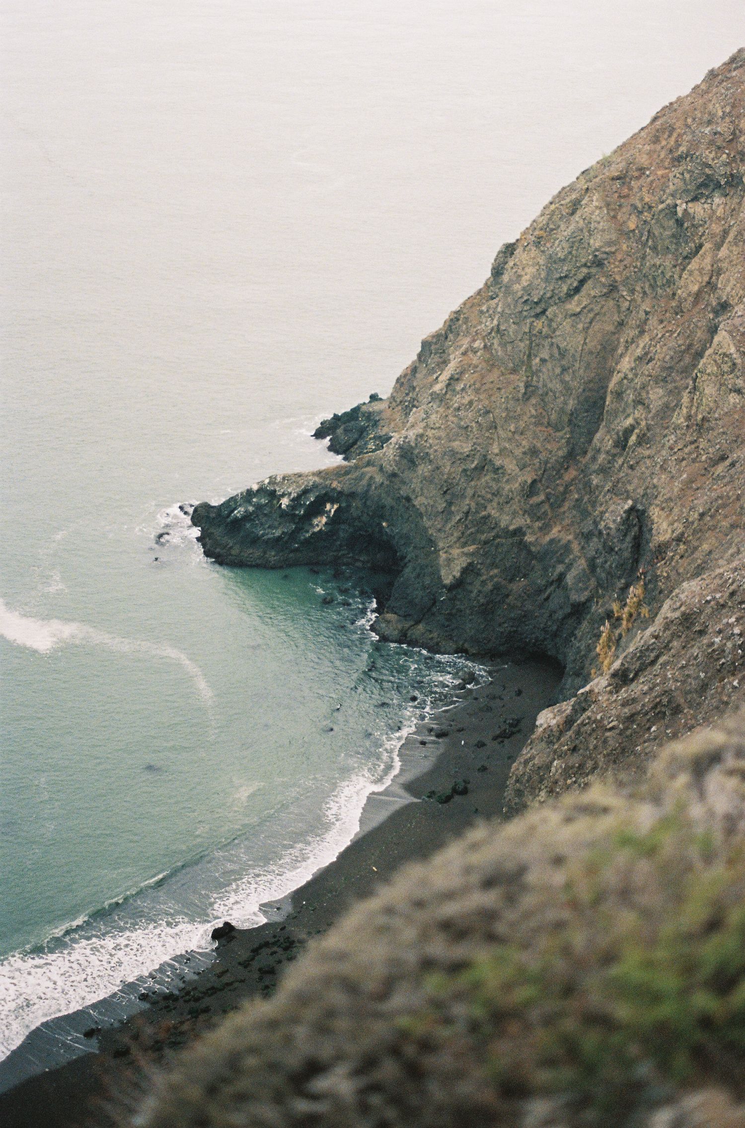 Aerial view of a rocky coastal cliff overlooking a secluded cove with turquoise waves and a dark sandy beach.