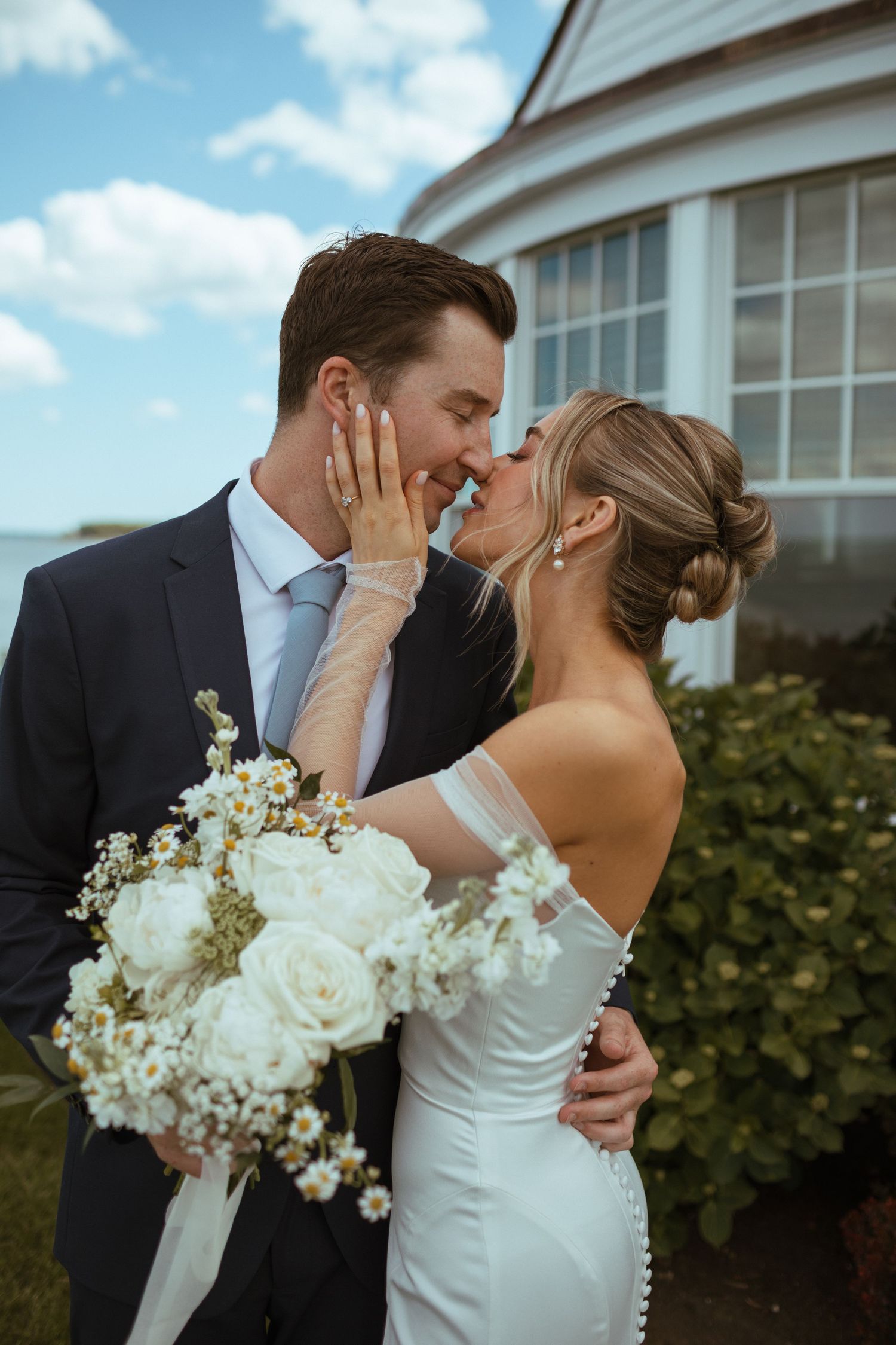 Bride and groom sharing a tender kiss outdoors, bride holding white rose bouquet in elegant wedding attire.