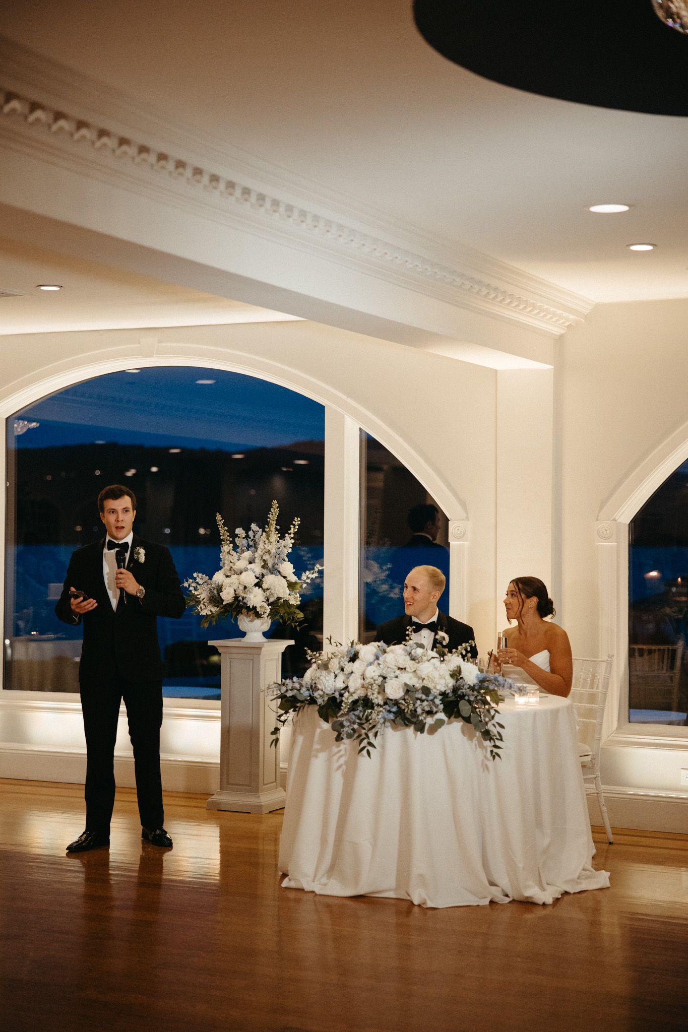 Best man giving a wedding toast while bride and groom sit at sweetheart table.