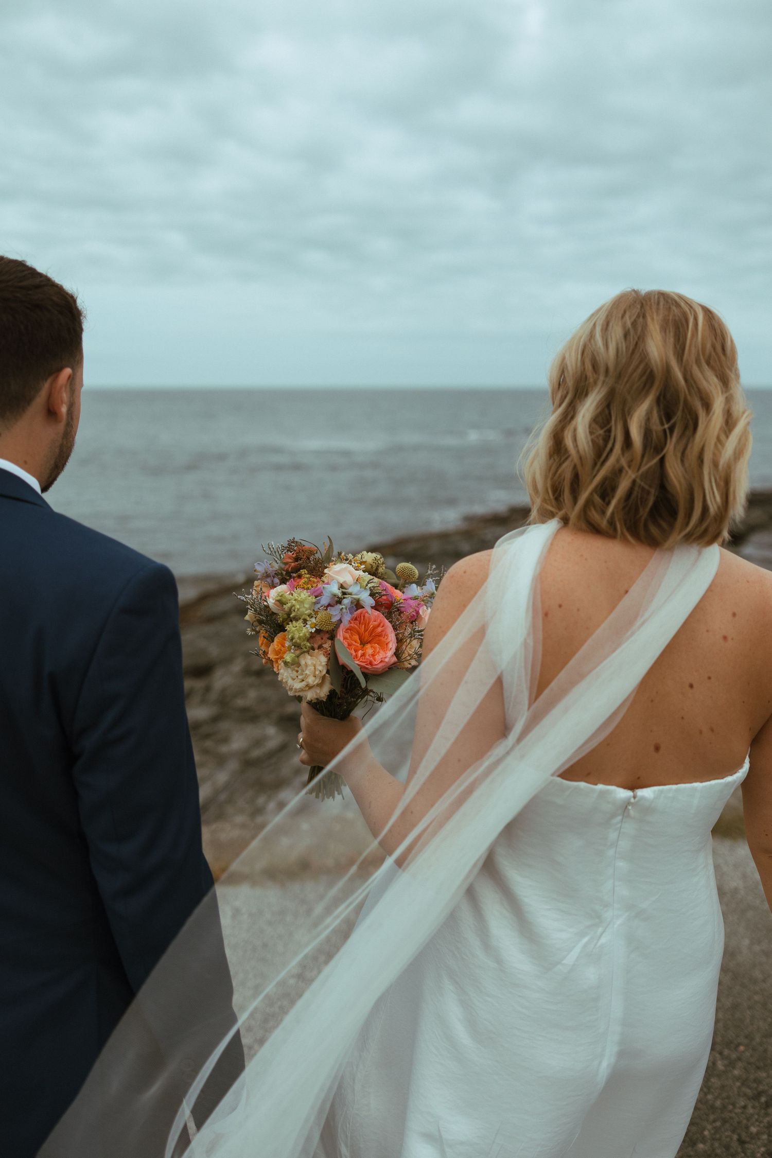 Bride and groom stand with backs to camera overlooking the rocky ocean, her veil and bouquet flowing in the breeze.